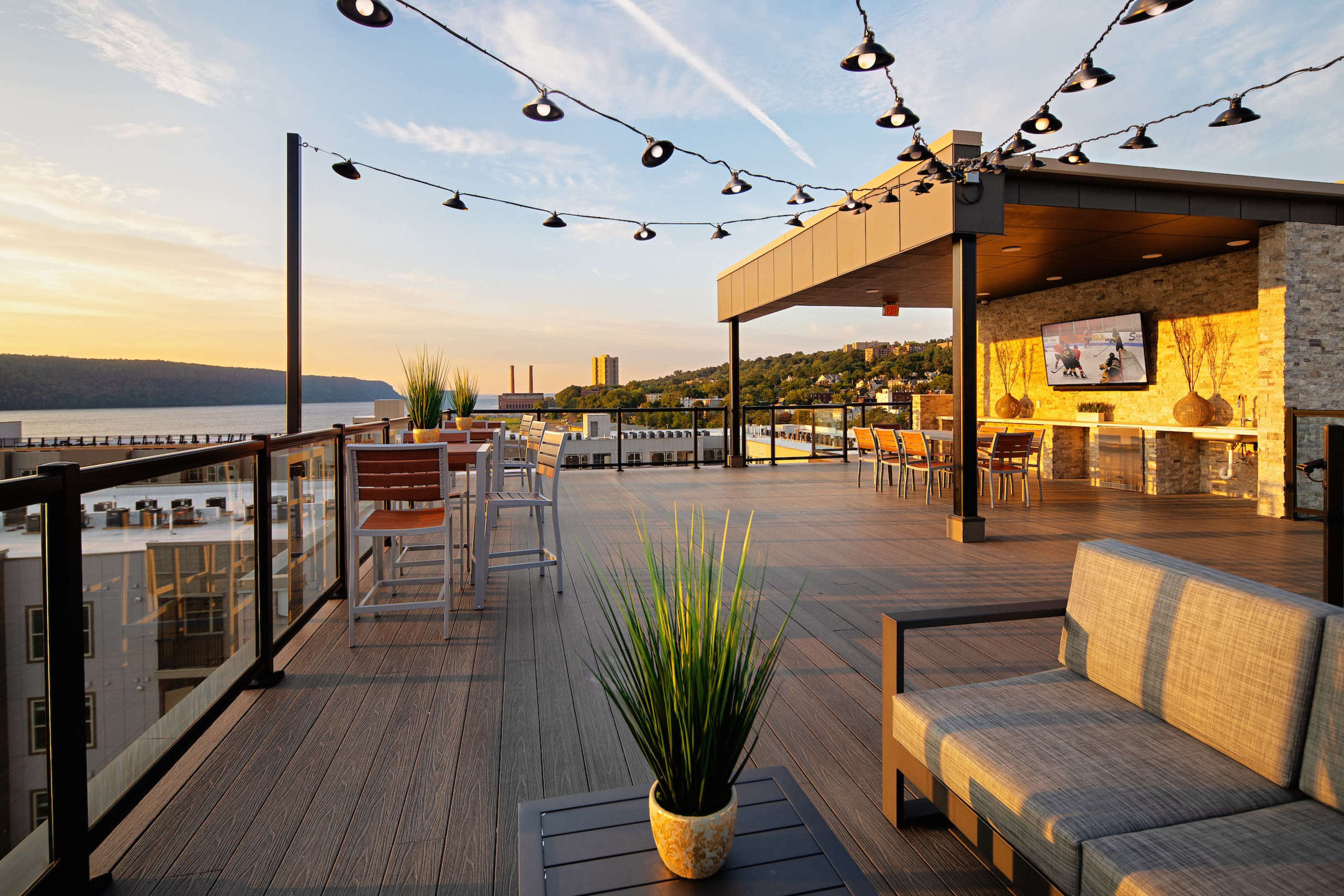 The image shows a rooftop terrace featuring modern seating, string lights, and a view of a landscape with buildings and a river at sunset.