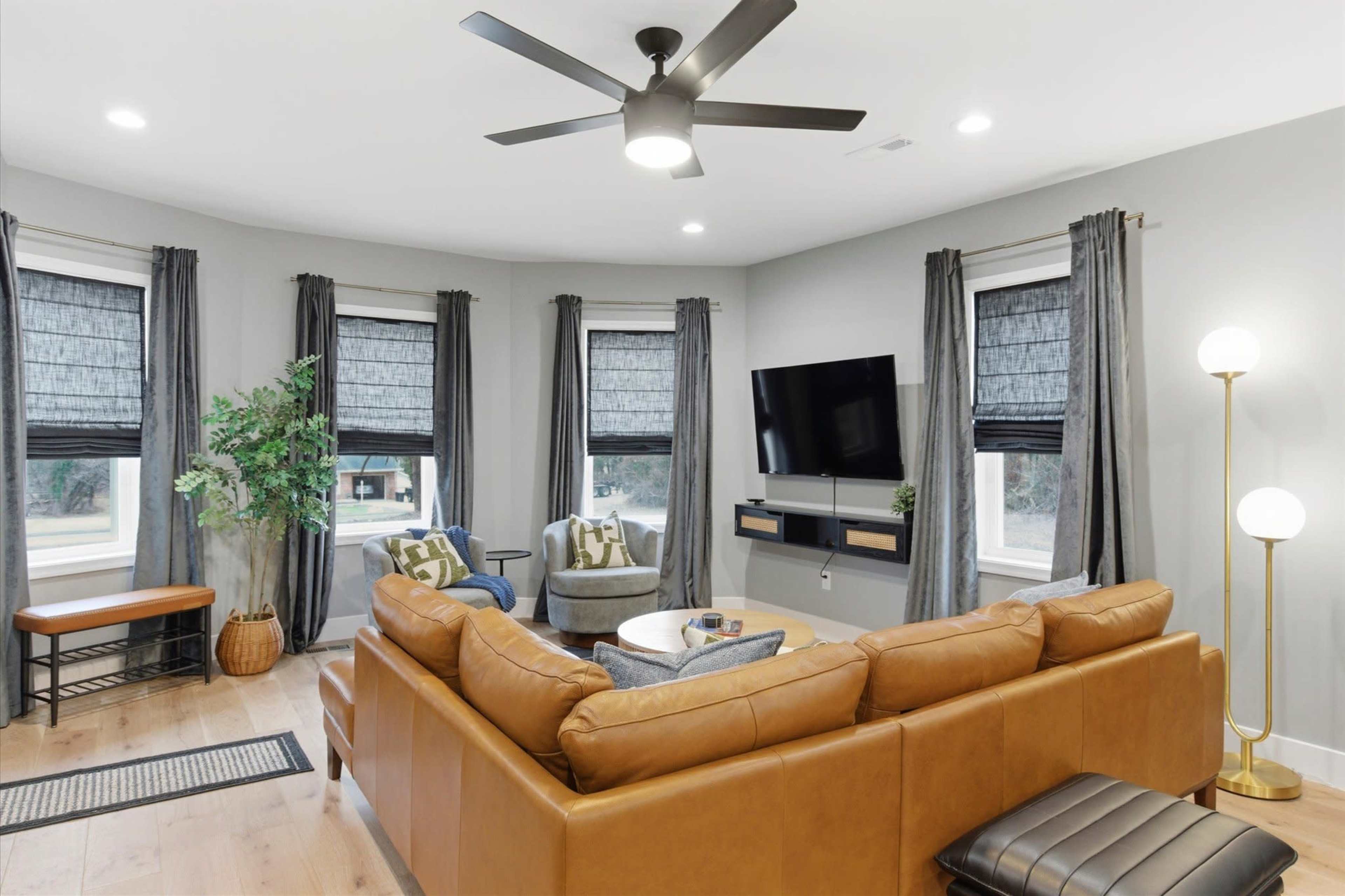 The image shows a modern living room with a large brown sectional sofa, a television mounted on the wall, and large windows with dark curtains, complemented by a ceiling fan and a light fixture.
