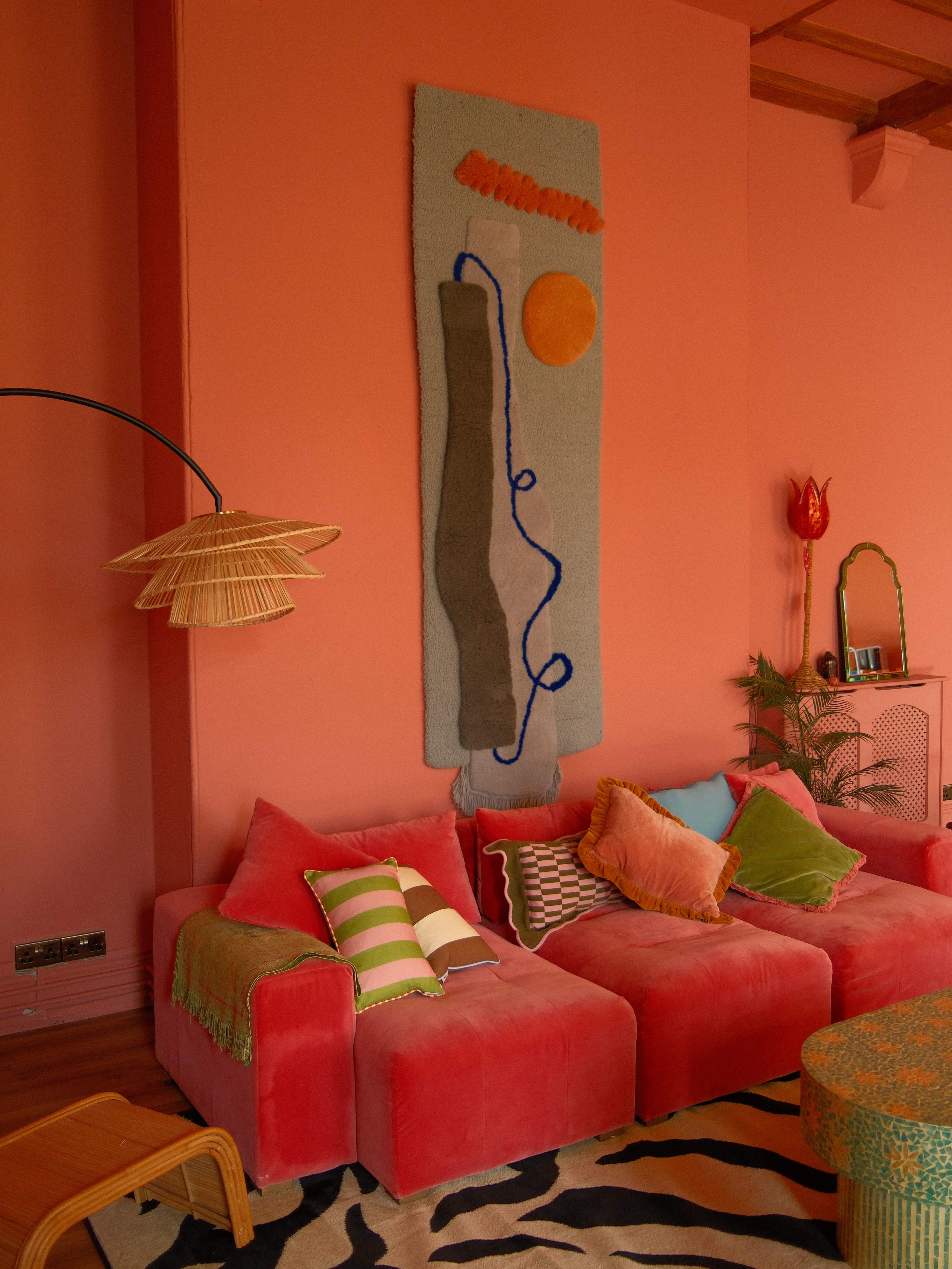 The image shows a vibrant living room with a large pink wall, a colorful abstract wall hanging, a sectional couch adorned with various cushions, and a patterned rug.