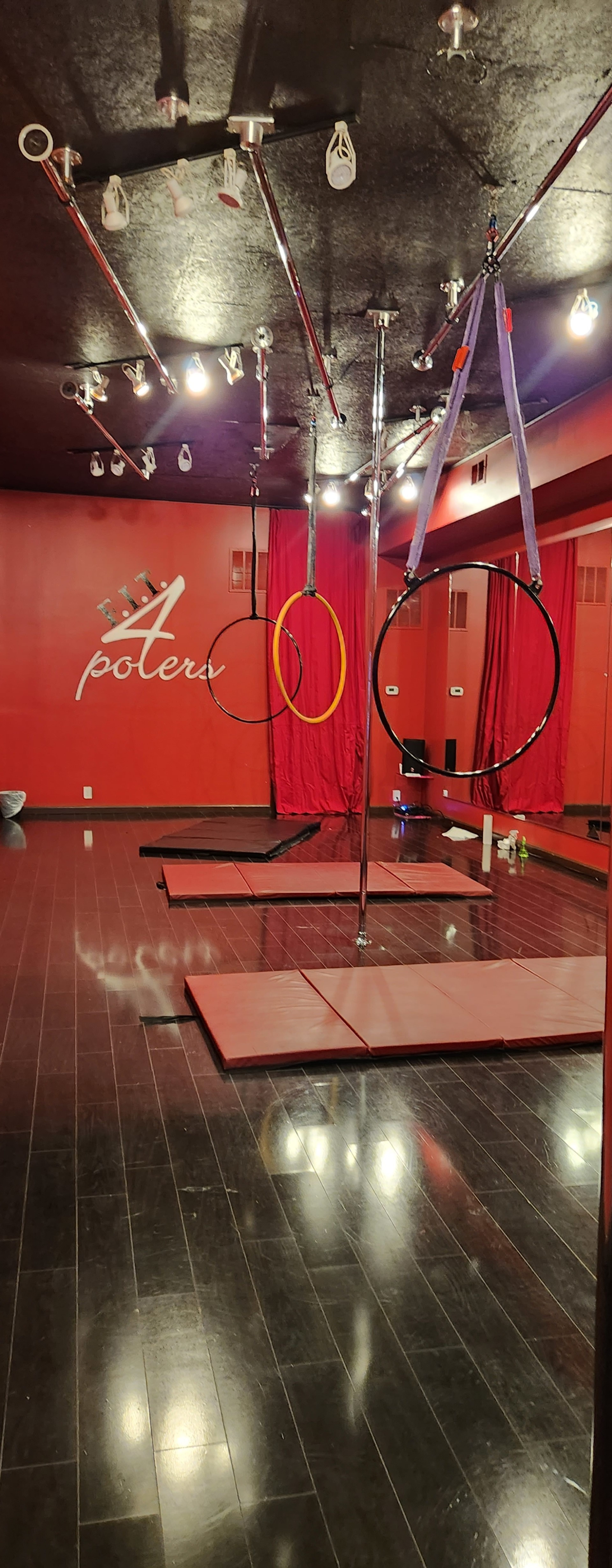 An empty aerial studio featuring a red wall with the word "poteru," two large aerial hoops suspended from the ceiling, and several exercise mats on the floor.