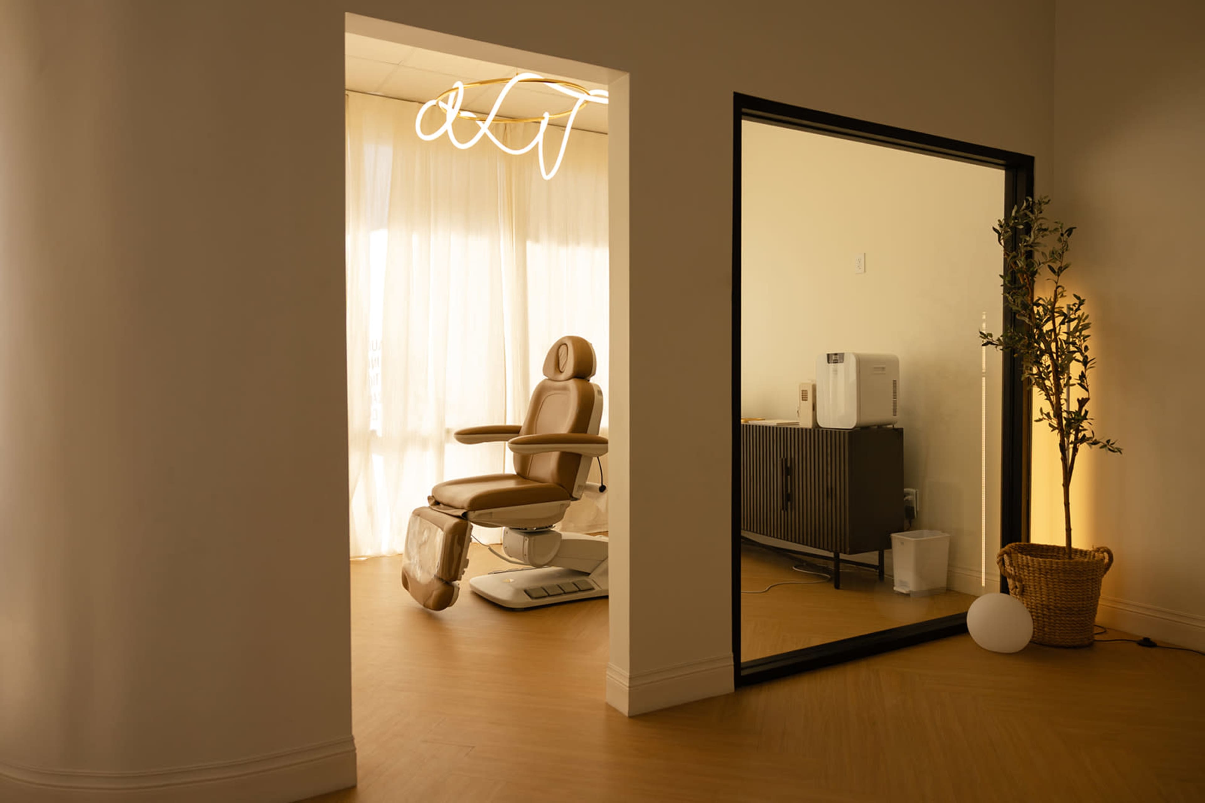 The image shows a minimalist interior space with a treatment chair visible through a doorway, alongside a dark cabinet and a potted plant in the corner.