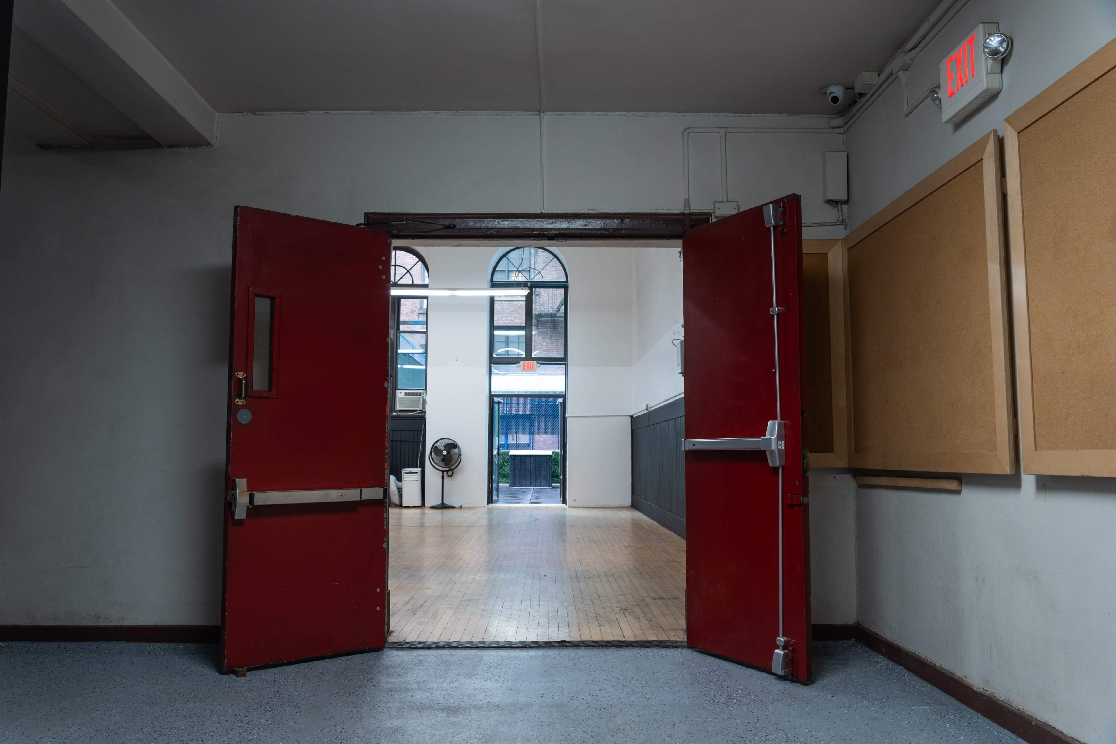 The image shows a pair of red double doors opening to a room with wooden flooring and large windows at the far end.
