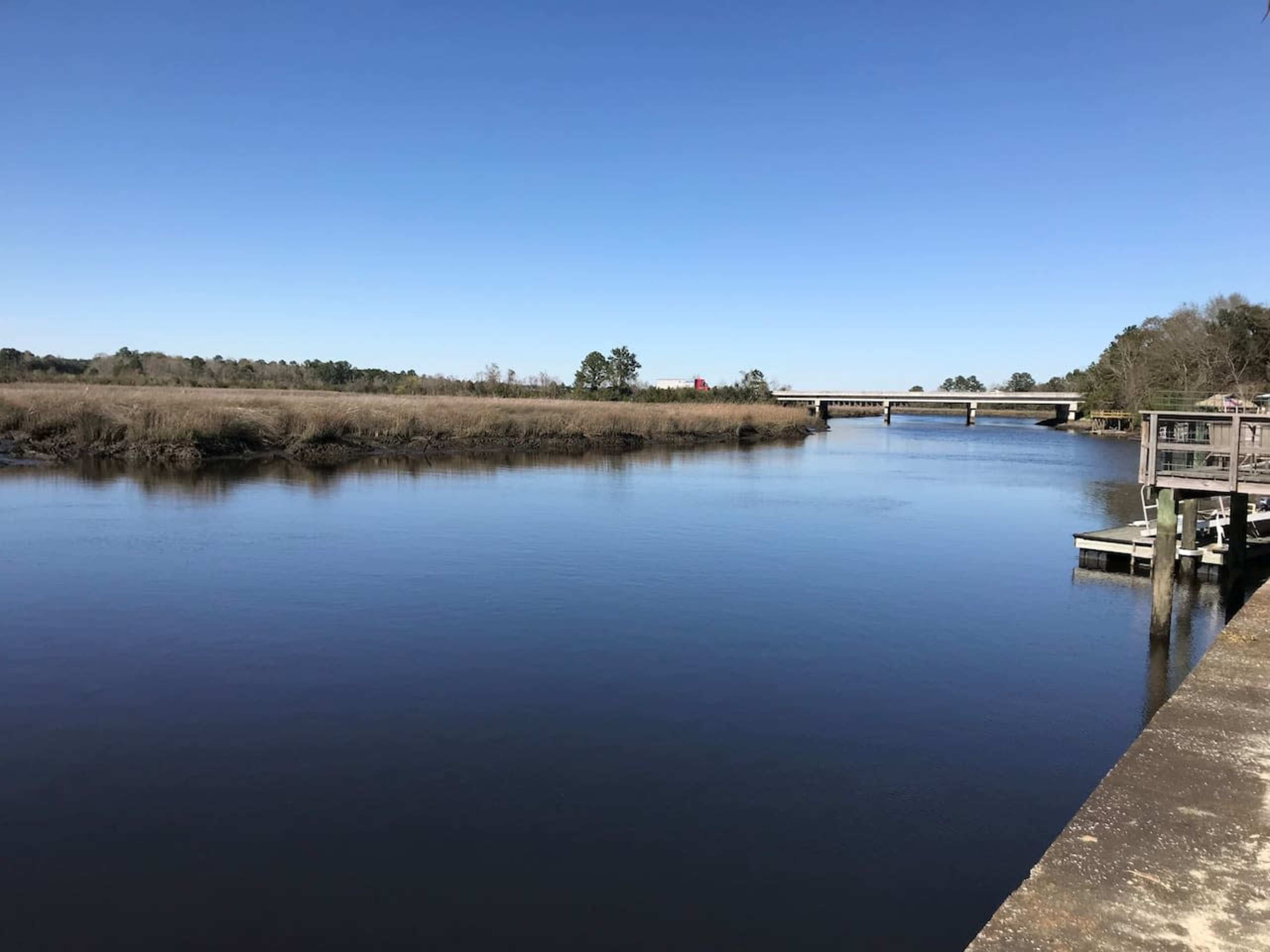 A calm river with a bridge crossing over it, surrounded by grassy banks and a clear blue sky.