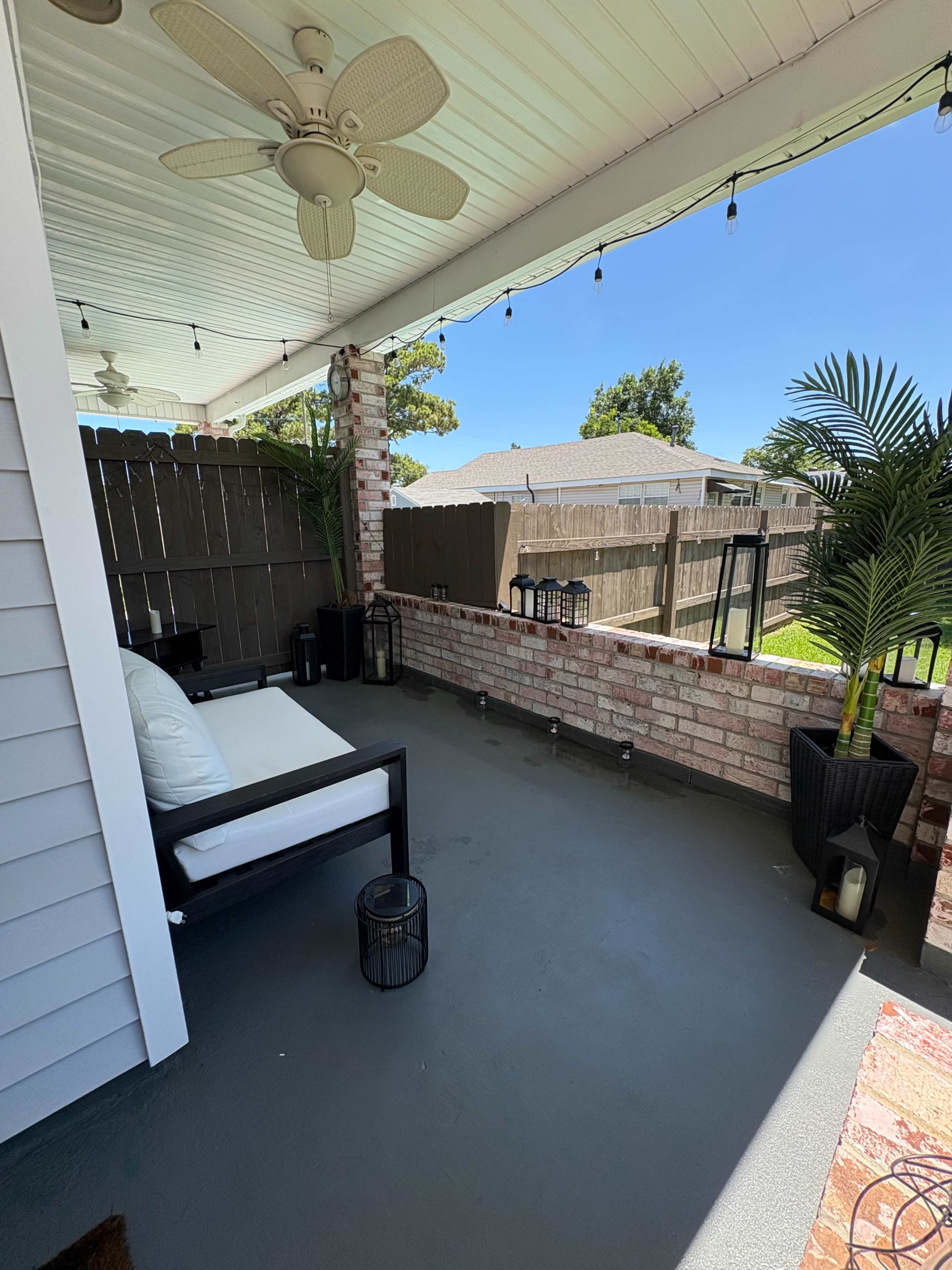 The image shows a covered patio area with a white couch, a ceiling fan, and potted plants, overlooking a fenced yard.