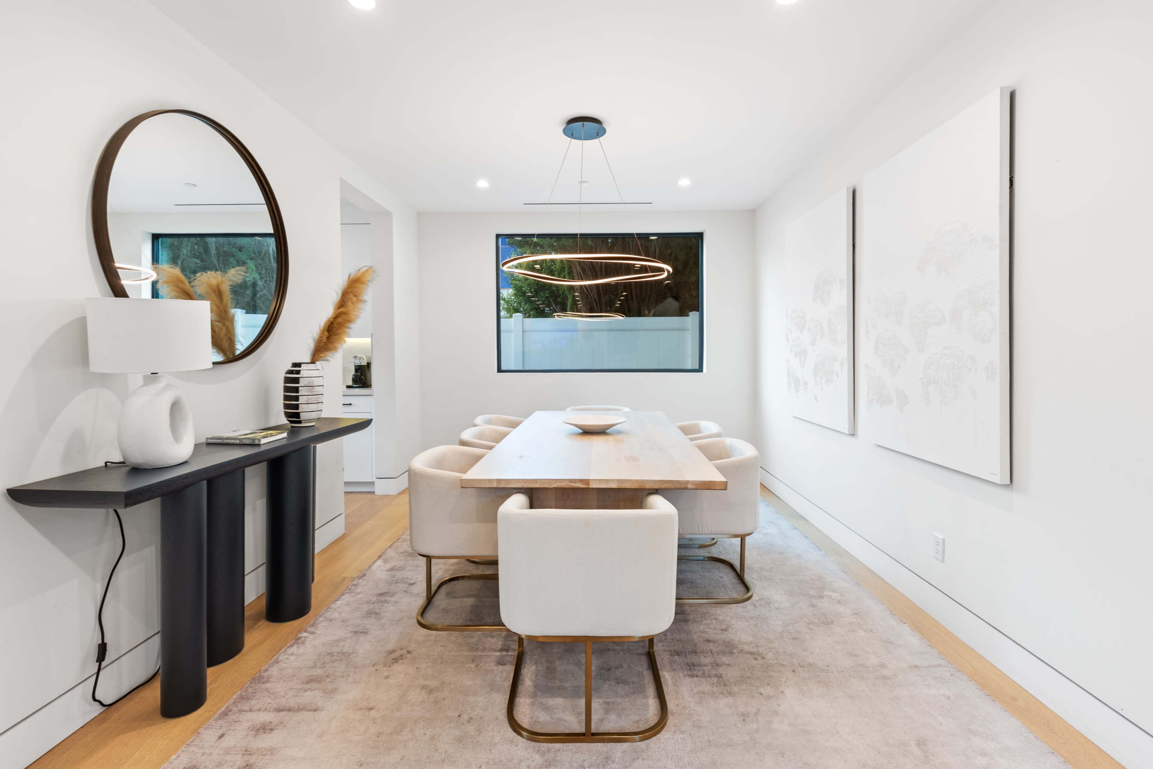 A modern dining room features a rectangular table surrounded by upholstered chairs, a circular mirror, and abstract wall art.