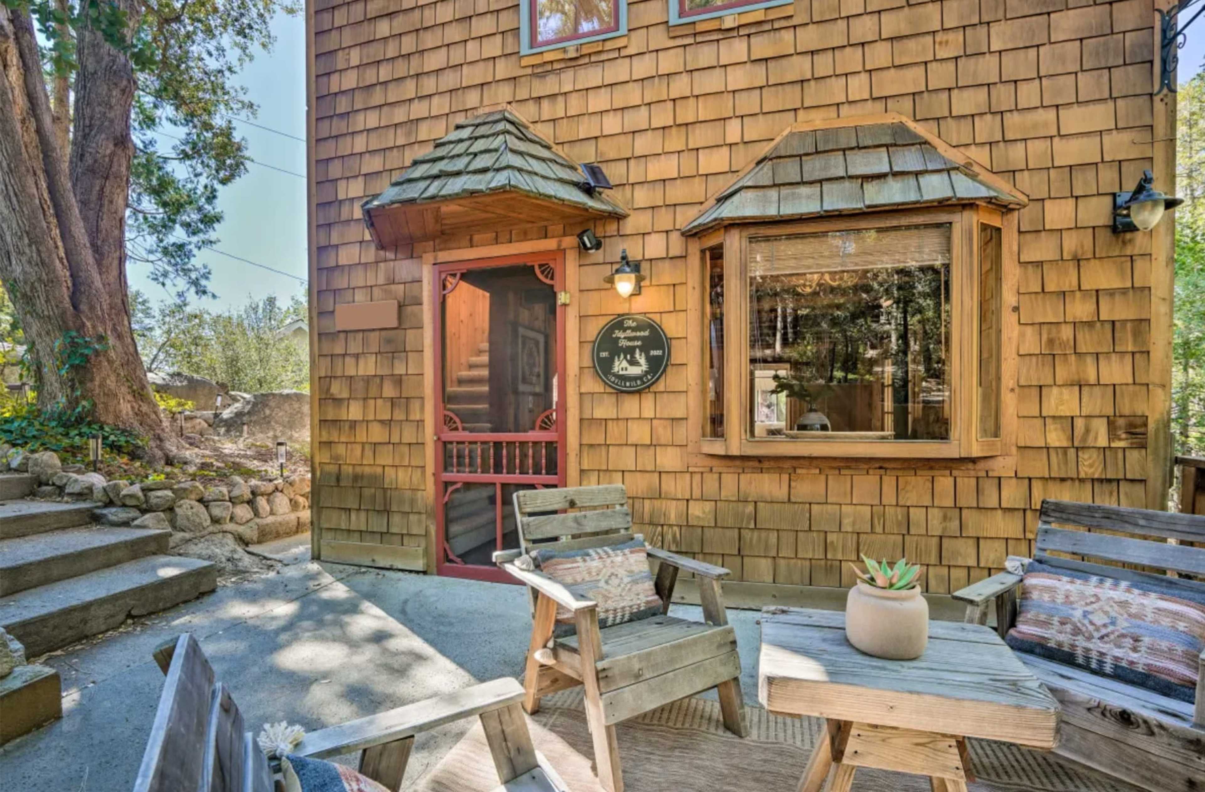 A wooden house with a shingled exterior, featuring a red door and a large window, accompanied by outdoor seating on a small patio.