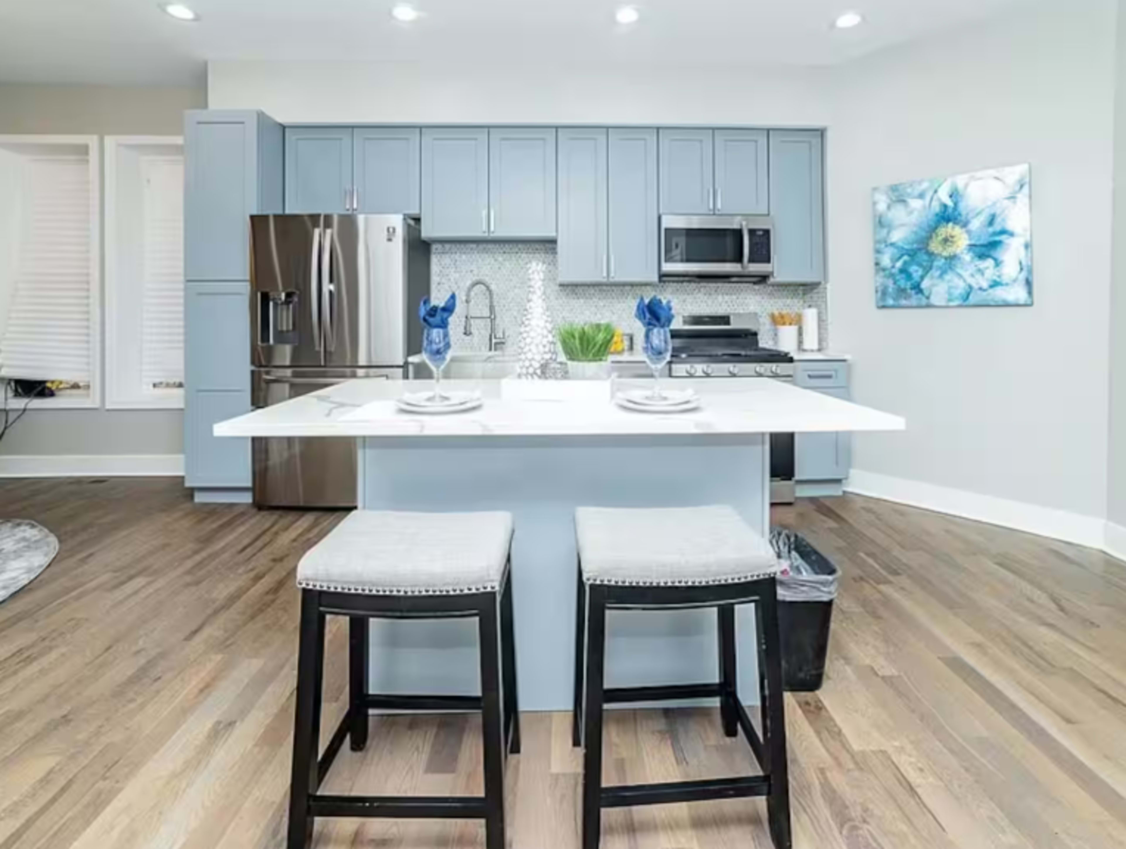 A modern kitchen features light blue cabinetry, stainless steel appliances, and a central island with two black stools.