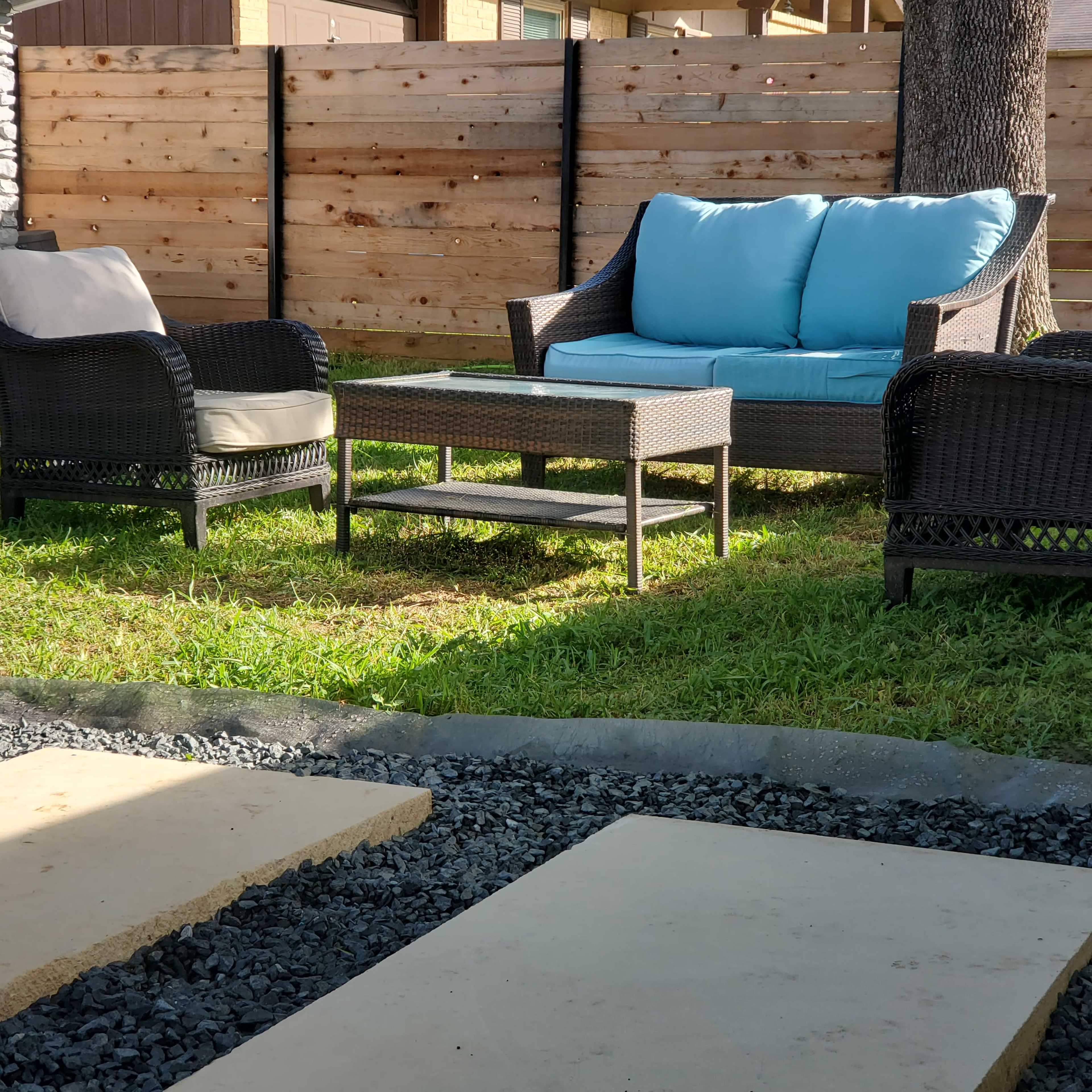 A patio area features two chairs and a couch with light blue cushions, accompanied by a glass-top table, all set on grass near a gravel pathway.