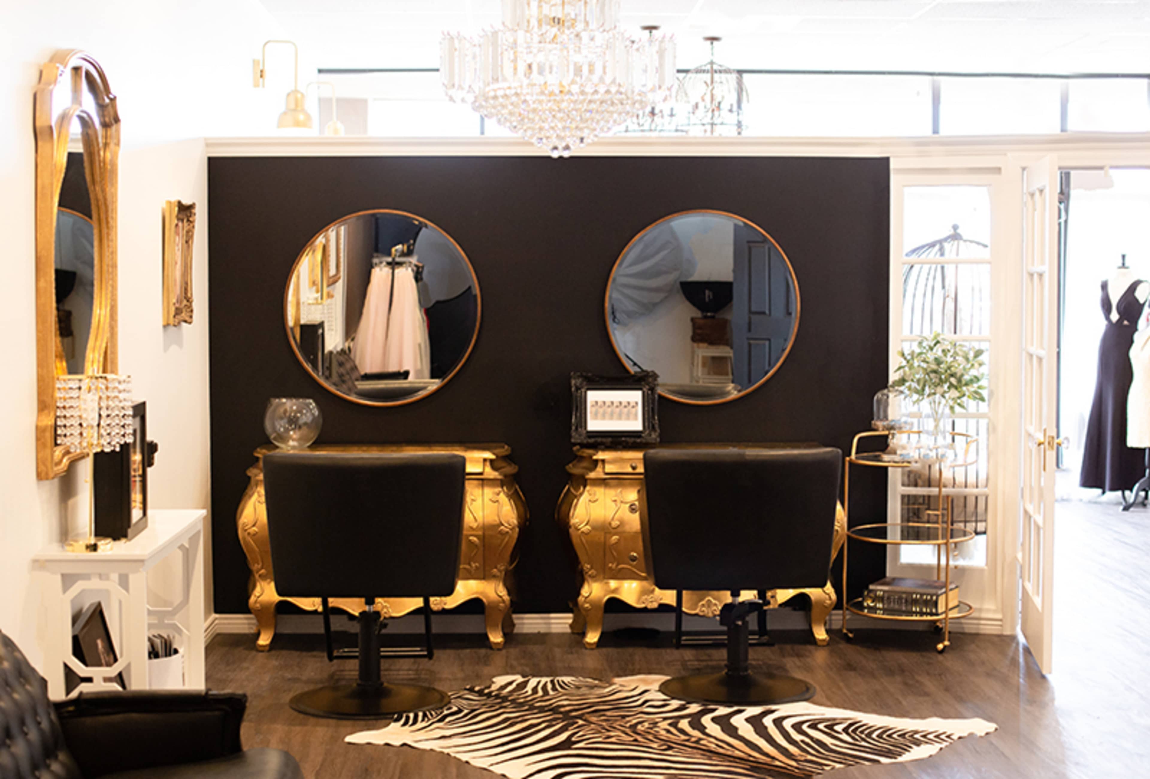 A stylish hair salon featuring two black chairs in front of golden vanity stations with round mirrors, set against a dark wall and adorned with a zebra print rug.
