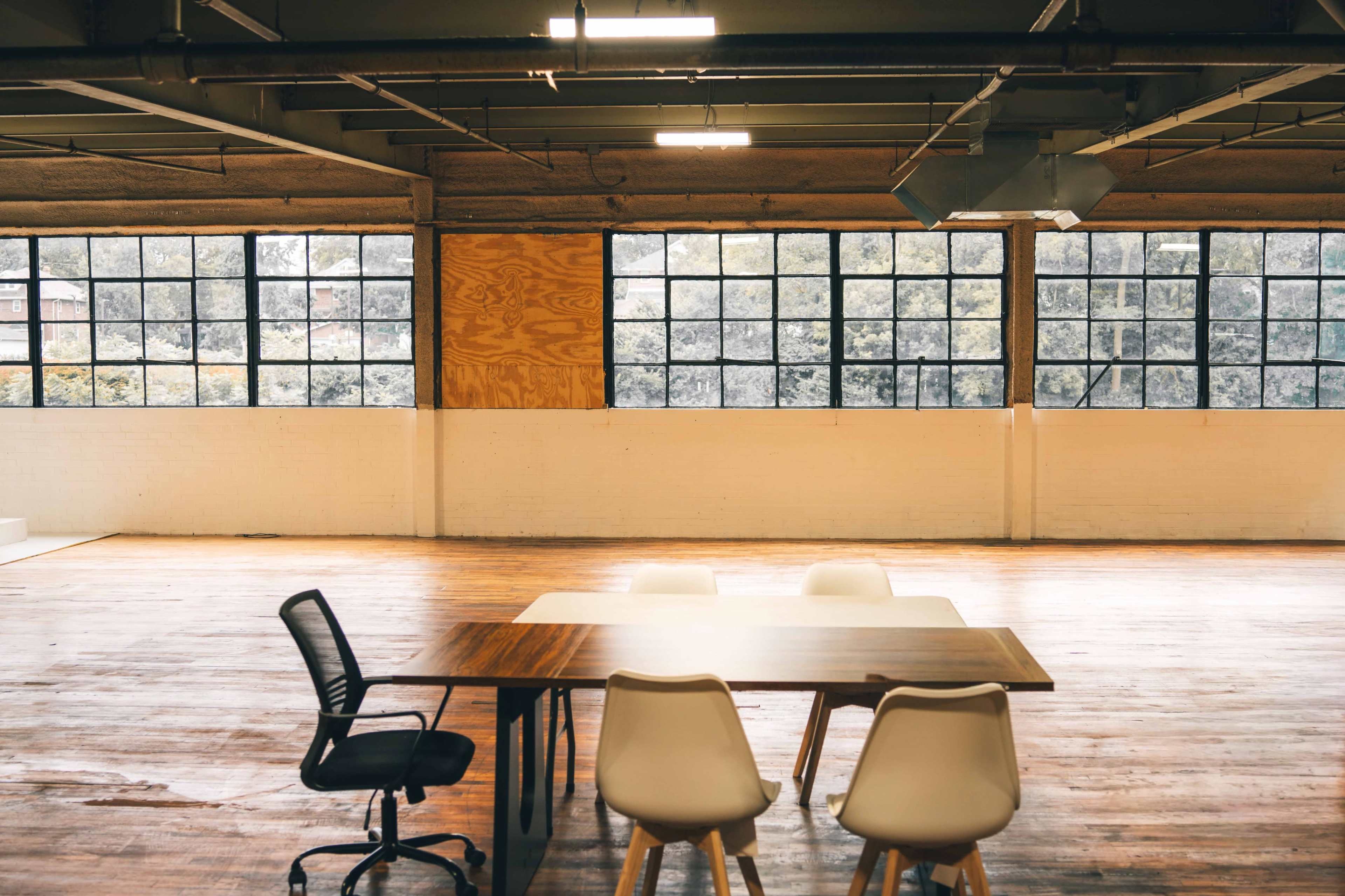 An empty industrial-style office space features a large wooden table with four white chairs and large windows that provide natural light.