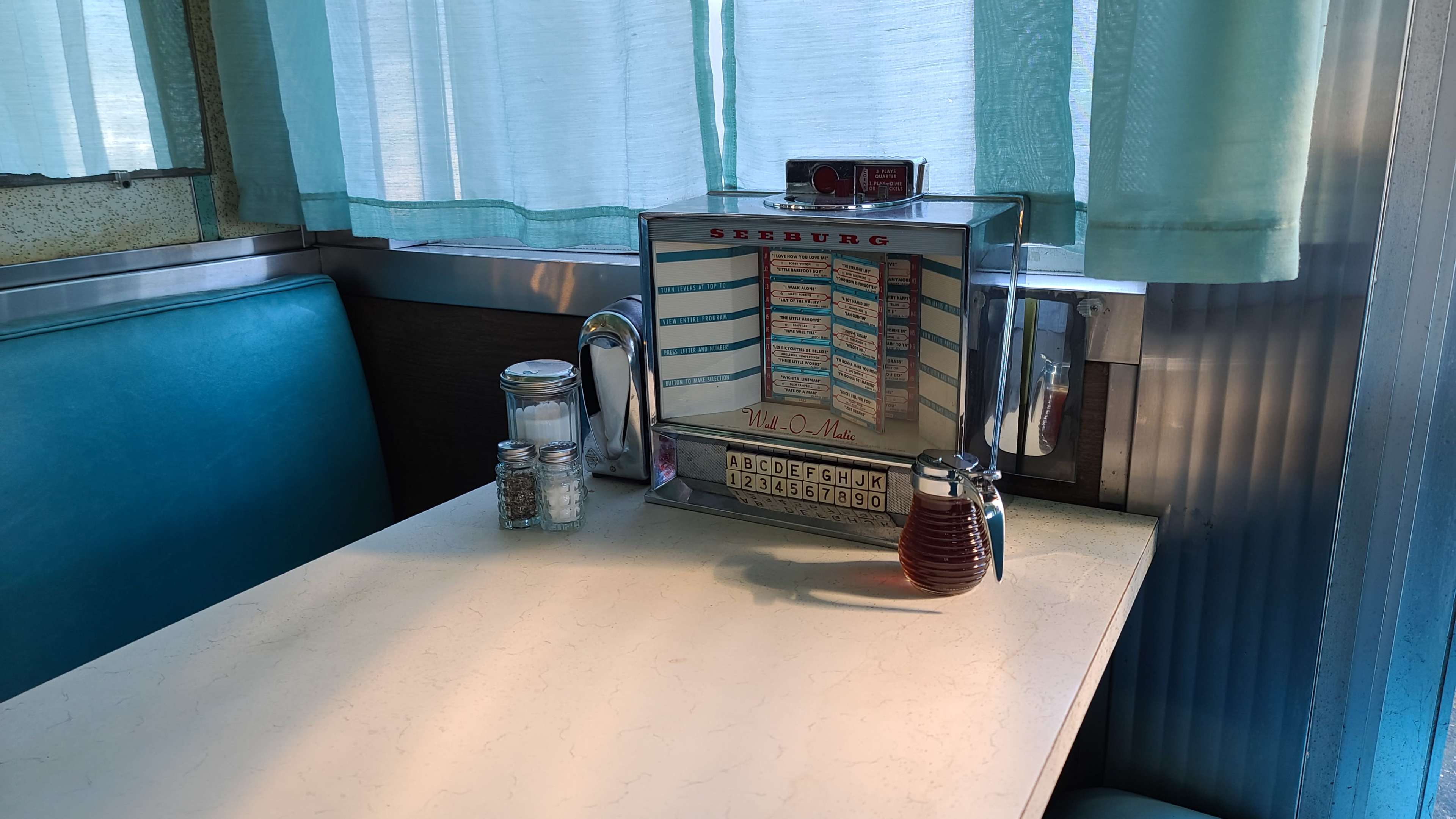 A diner table with a Seeburg jukebox, salt and pepper shakers, and a glass bottle of syrup, all beneath a window with light blue curtains.