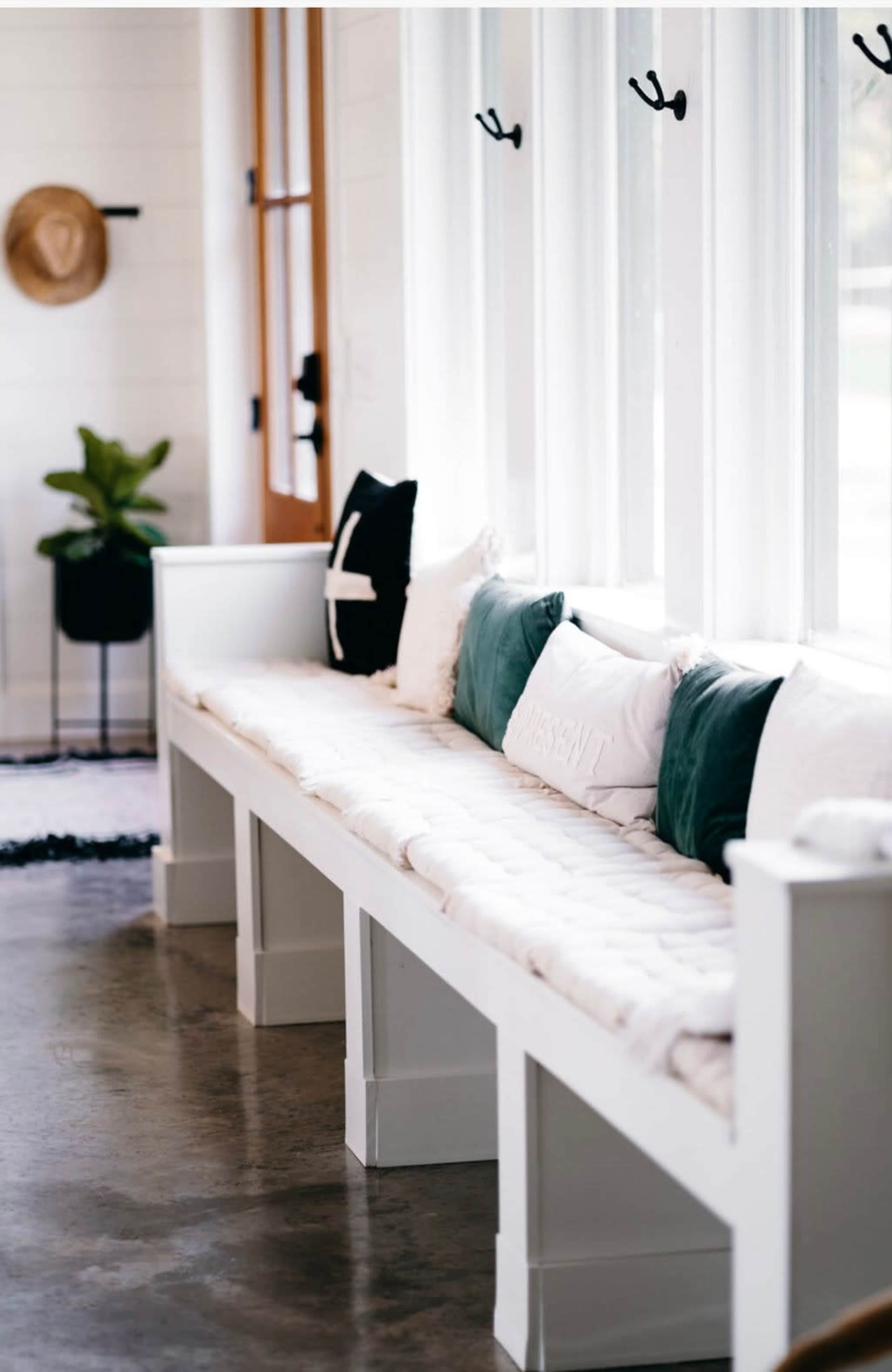 A long white bench with cushions and decorative pillows is positioned near large windows in a bright interior space.