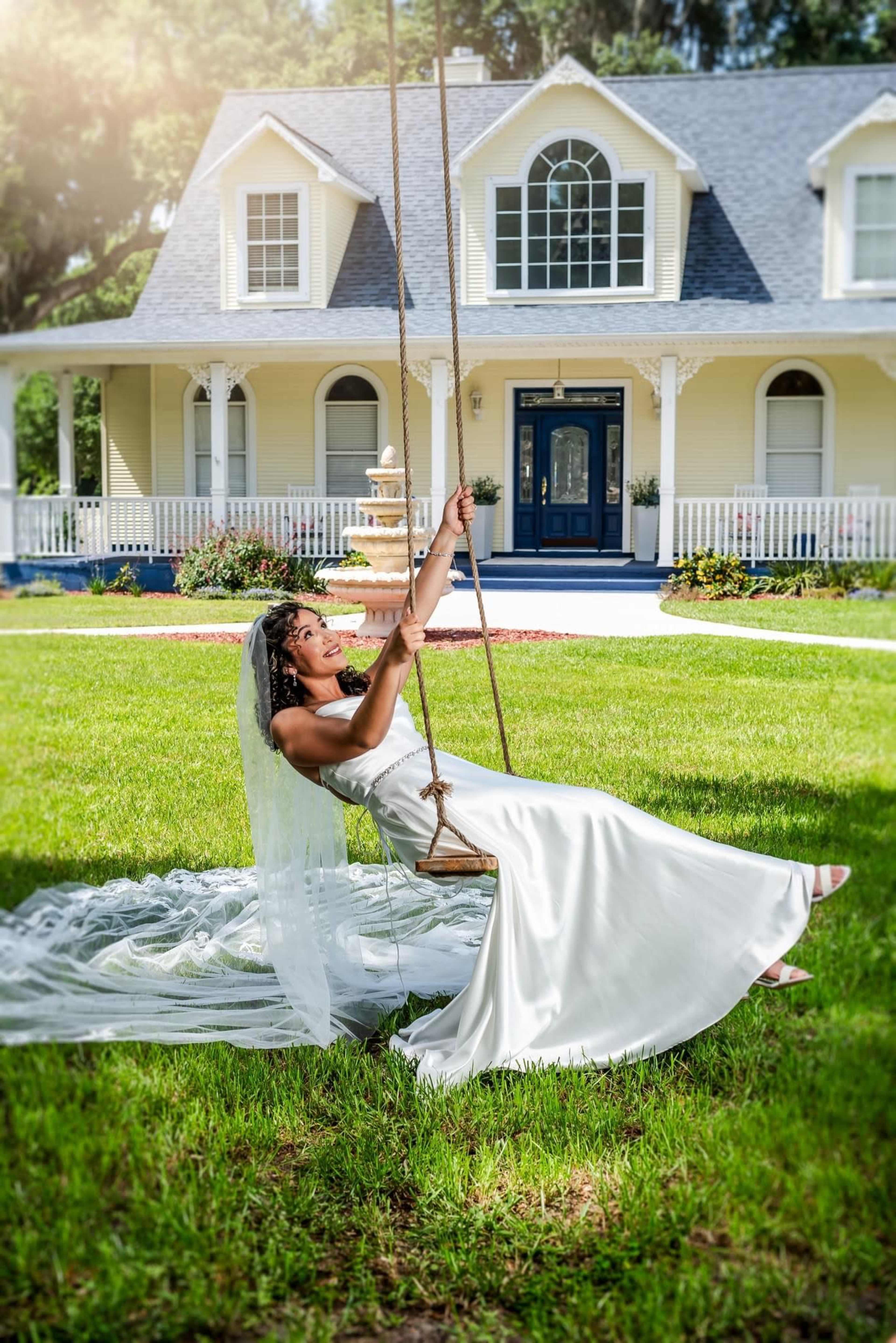A bride in a white gown swings on a rope swing in front of a large yellow house.