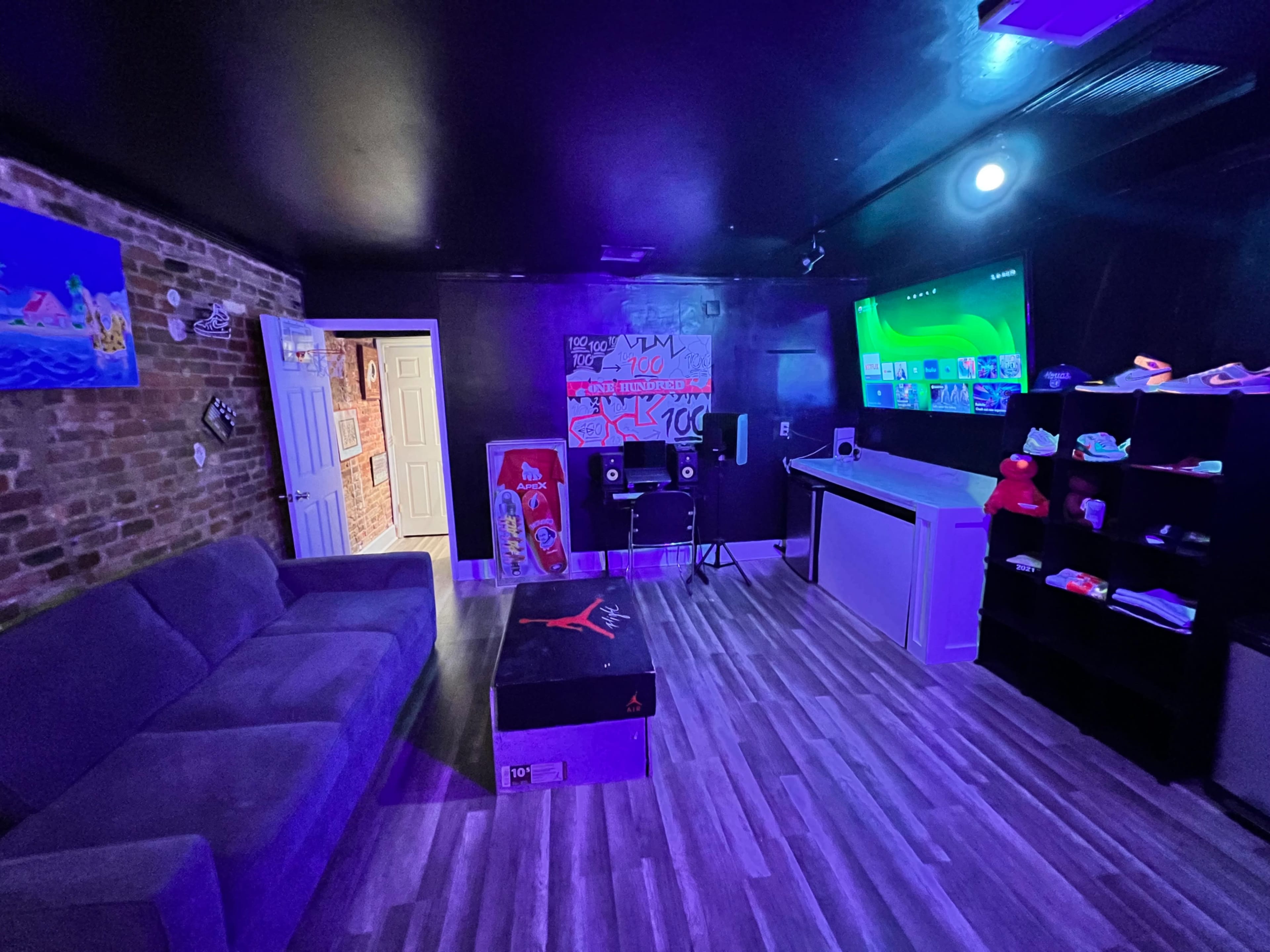The room features a darkly designed lounge area with a grey couch, a wall displaying colorful art, a large screen on one wall, and shelves holding various shoes.