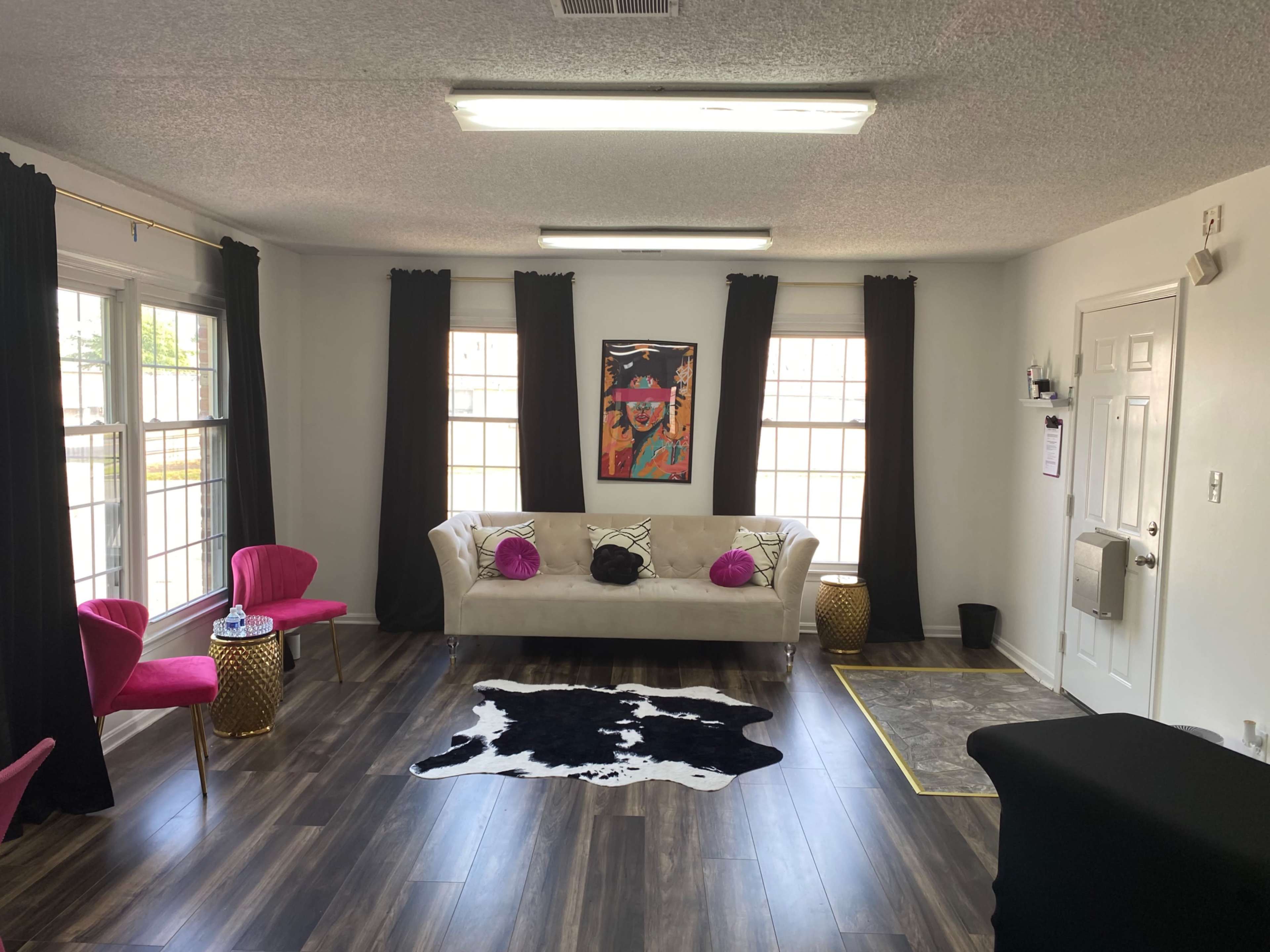 The room features a white sofa with black and pink accents, two pink chairs, a cowhide rug, and large windows with black curtains.
