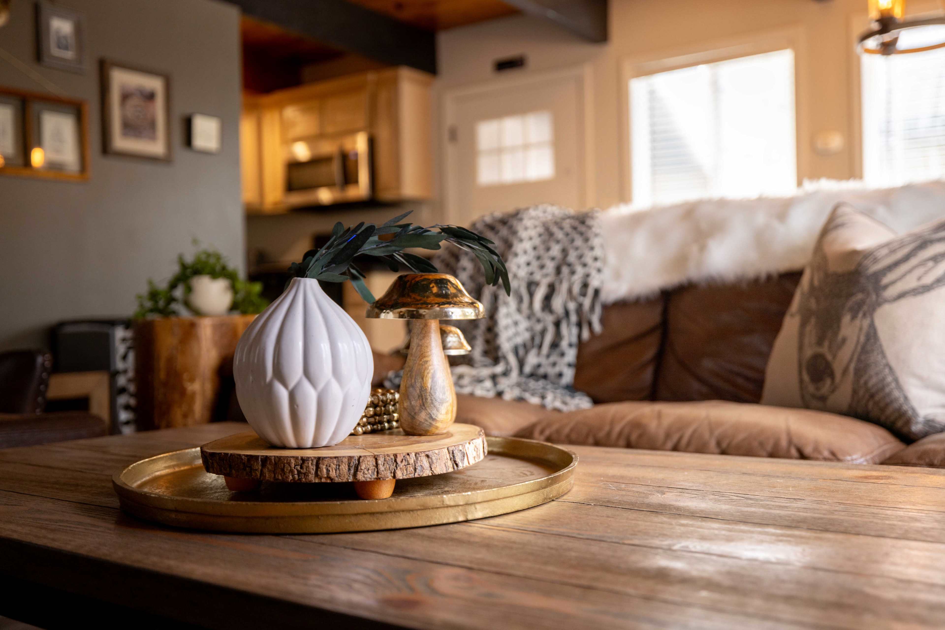 A living room scene with a wooden coffee table displaying a white vase, a decorative mushroom figure, and a wooden tray, set against a backdrop of a brown leather couch and a well-lit kitchen area.