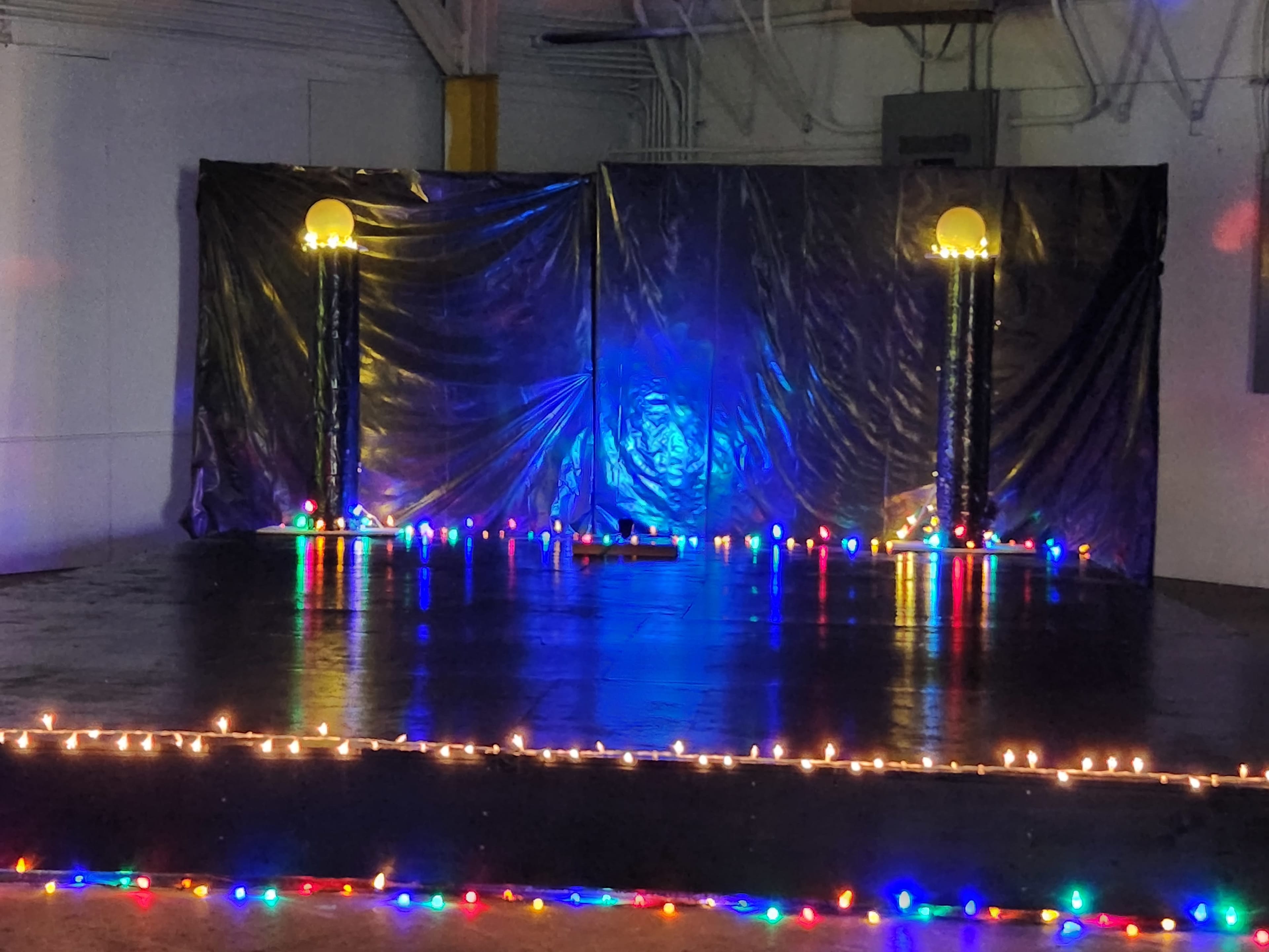The image shows a stage with a black backdrop and illuminated columns on either side, adorned with colorful lights along the floor.