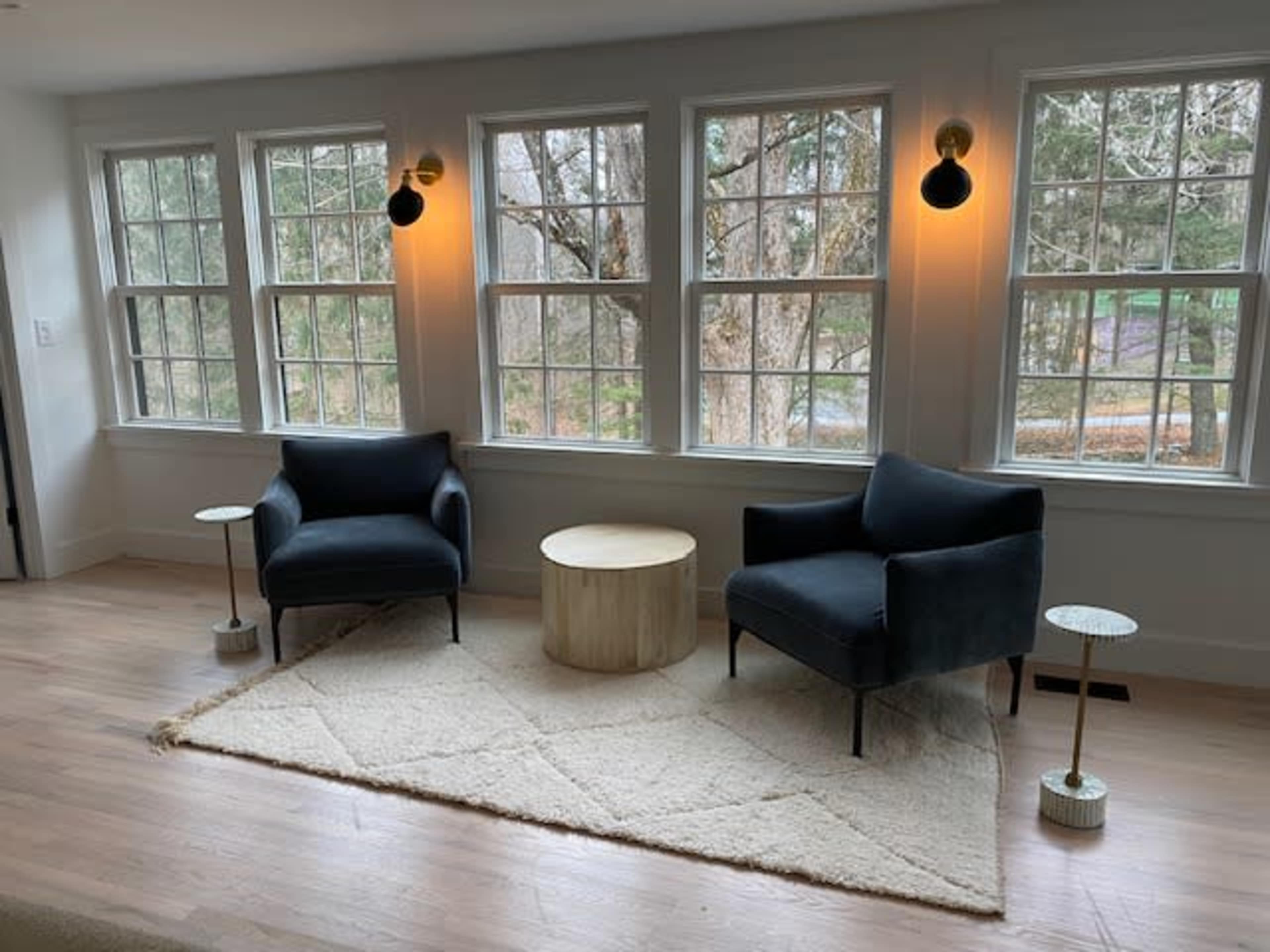 The room features two dark chairs and a round wooden table, set on a light rug, with large windows allowing natural light to illuminate the space.