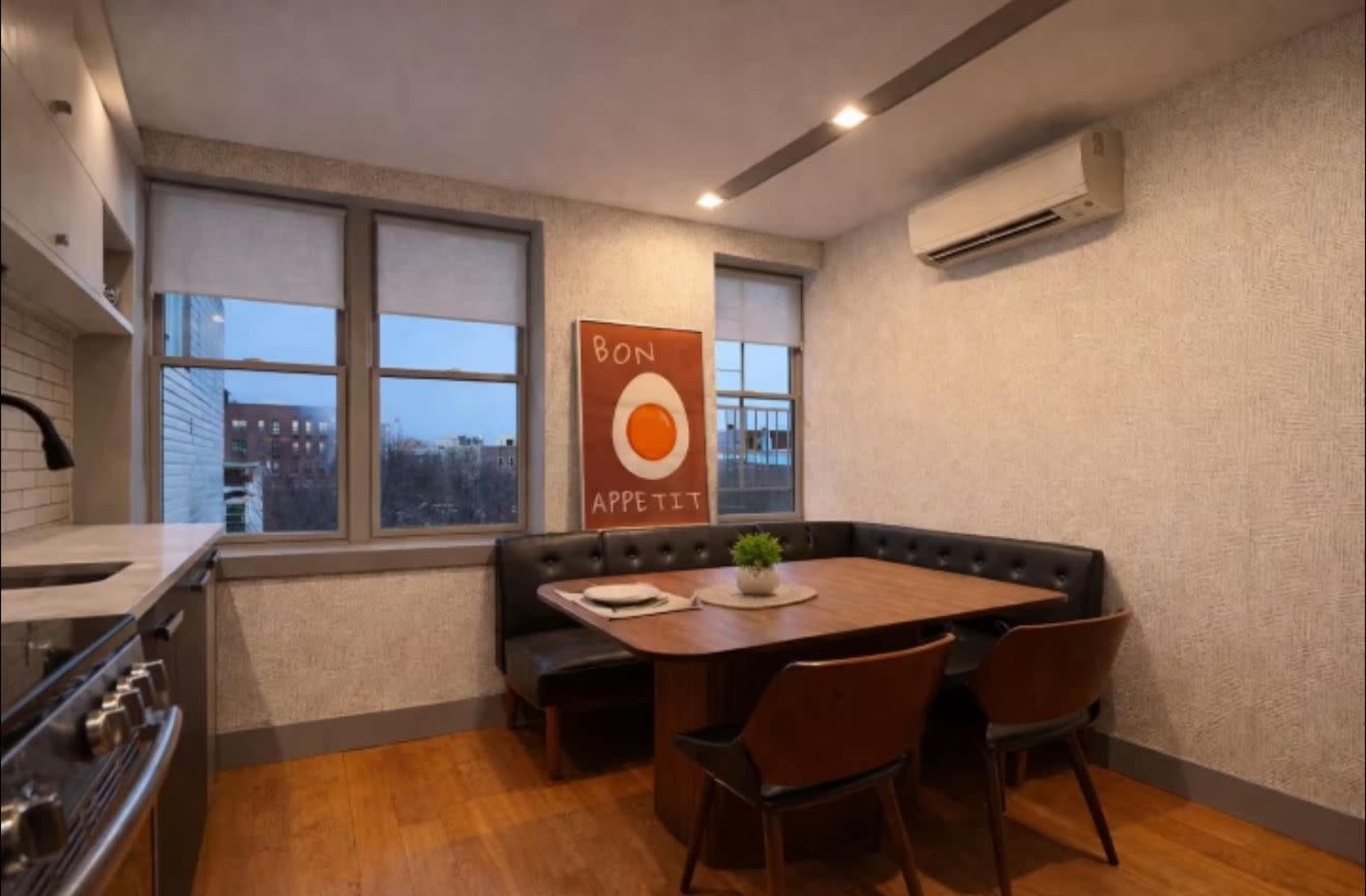 The image shows a modern kitchen with a dining nook featuring a wooden table and black chairs, flanked by windows and a decorative wall art piece.