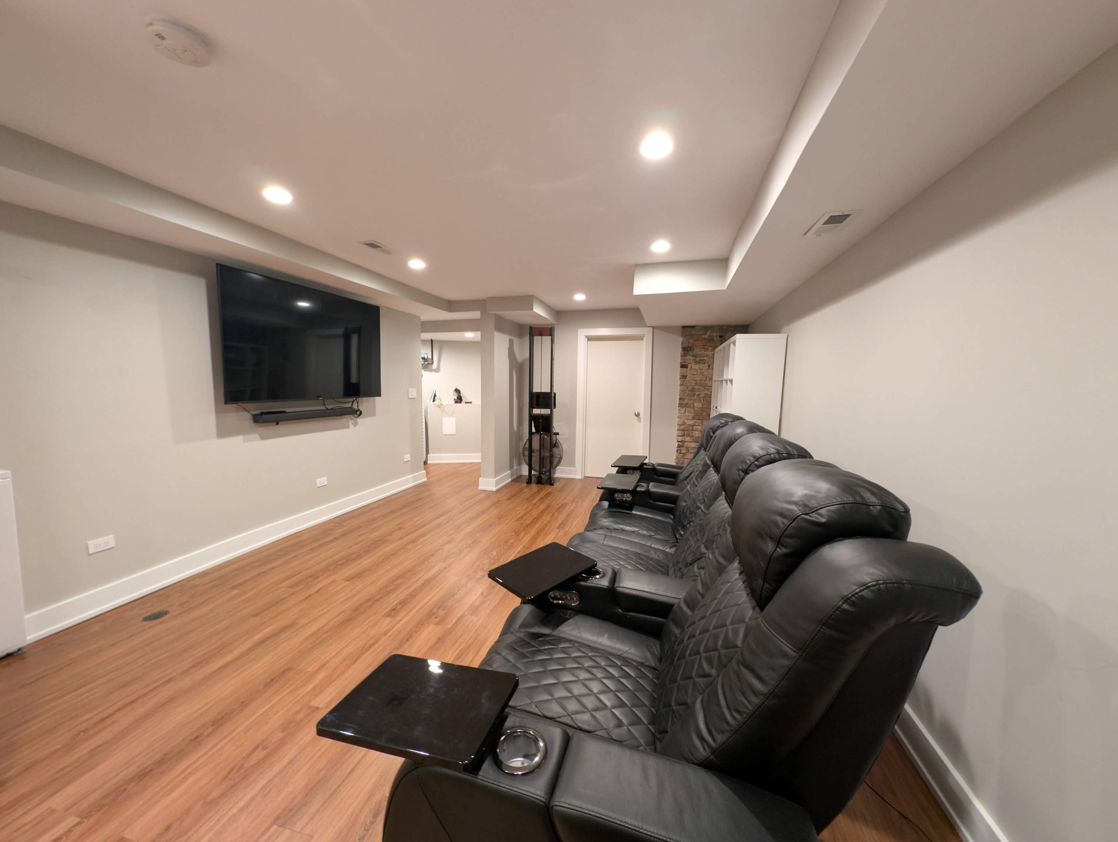 The image shows a modern entertainment room with black reclining chairs facing a flat-screen TV mounted on the wall.