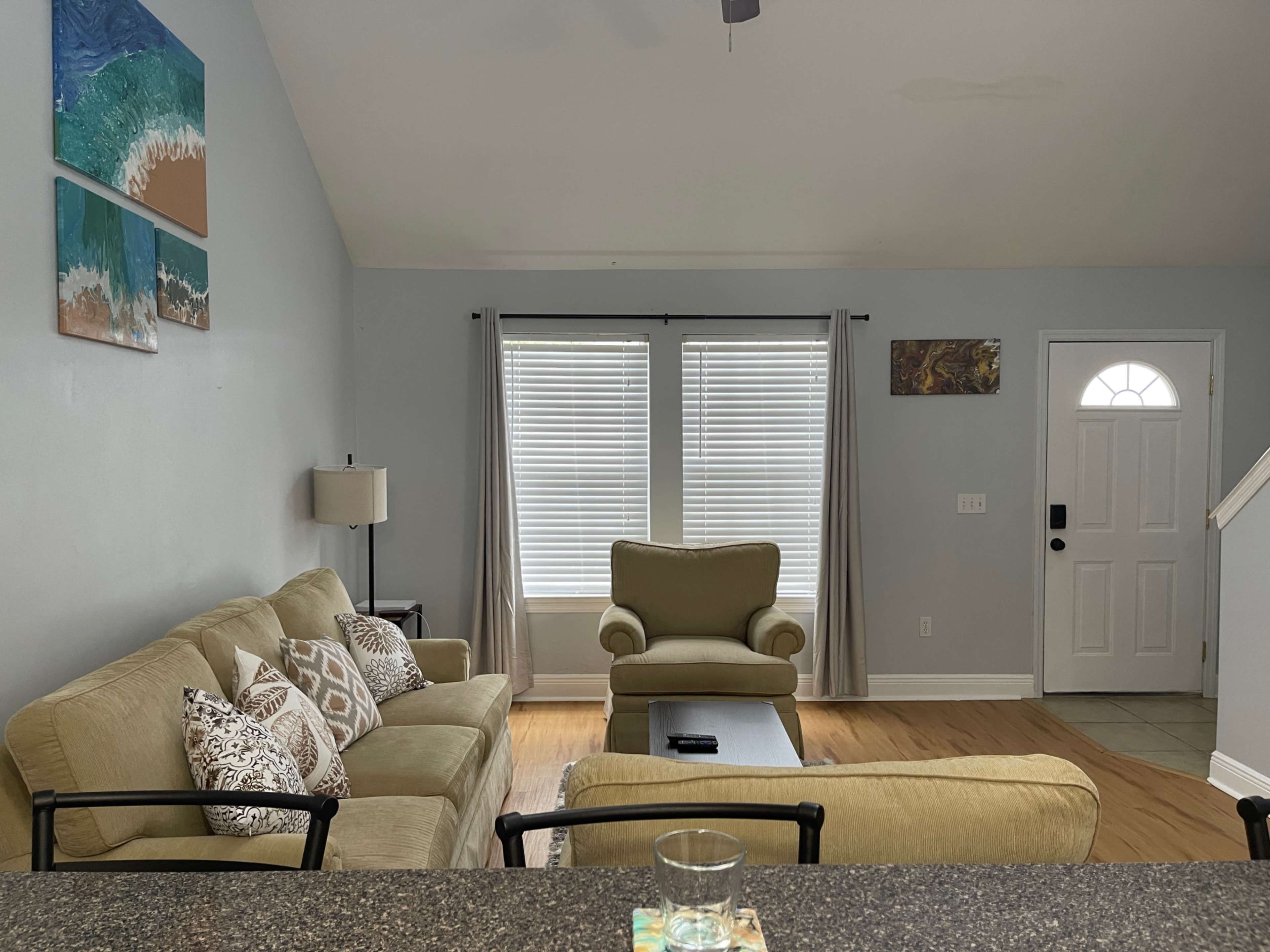 A living room with a light green couch and armchair, large windows with blinds, and a door leading outside.