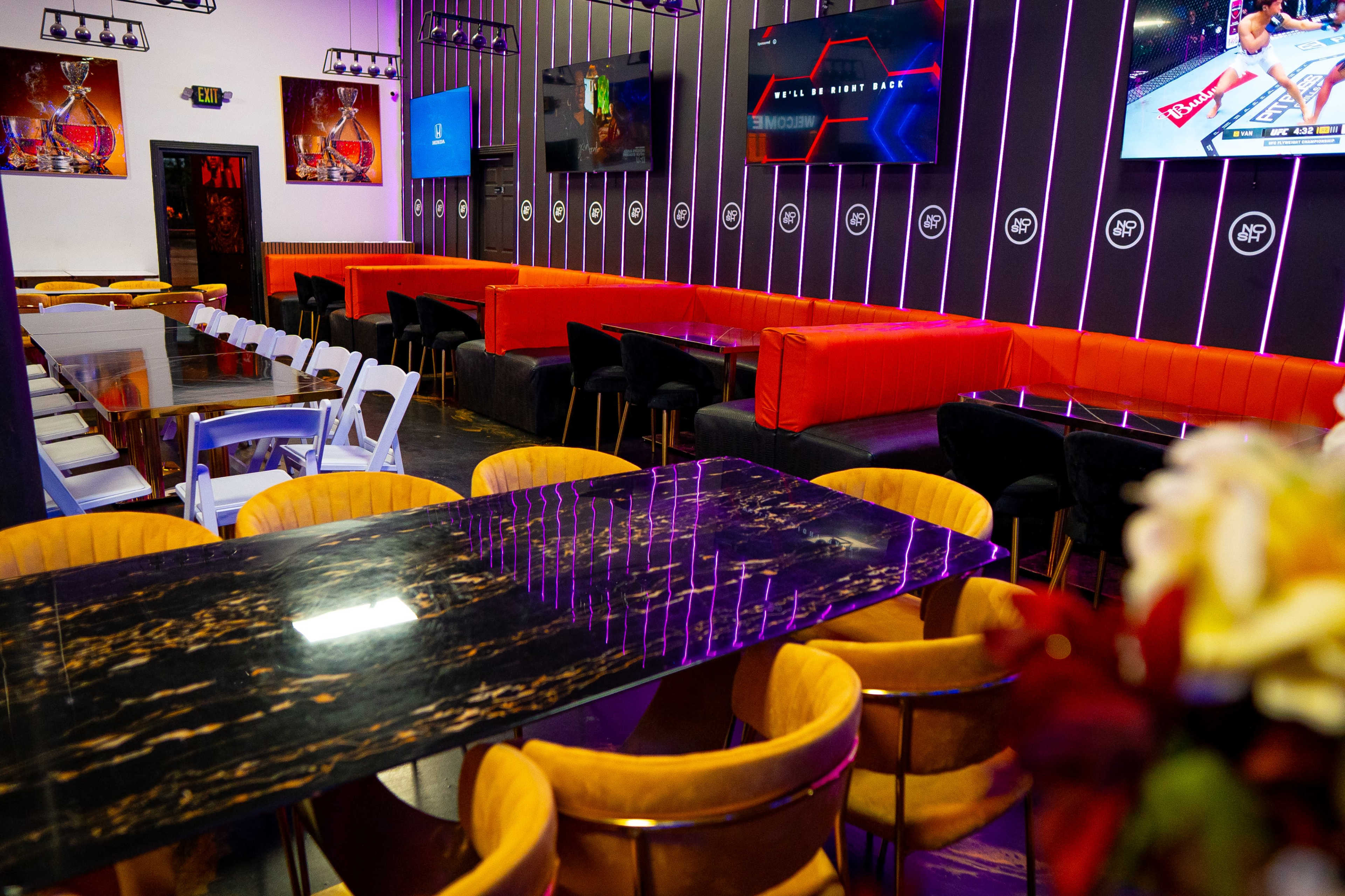 The image shows a modern dining area with a mix of colorful seating, marble tables, and wall-mounted televisions displaying sports events.