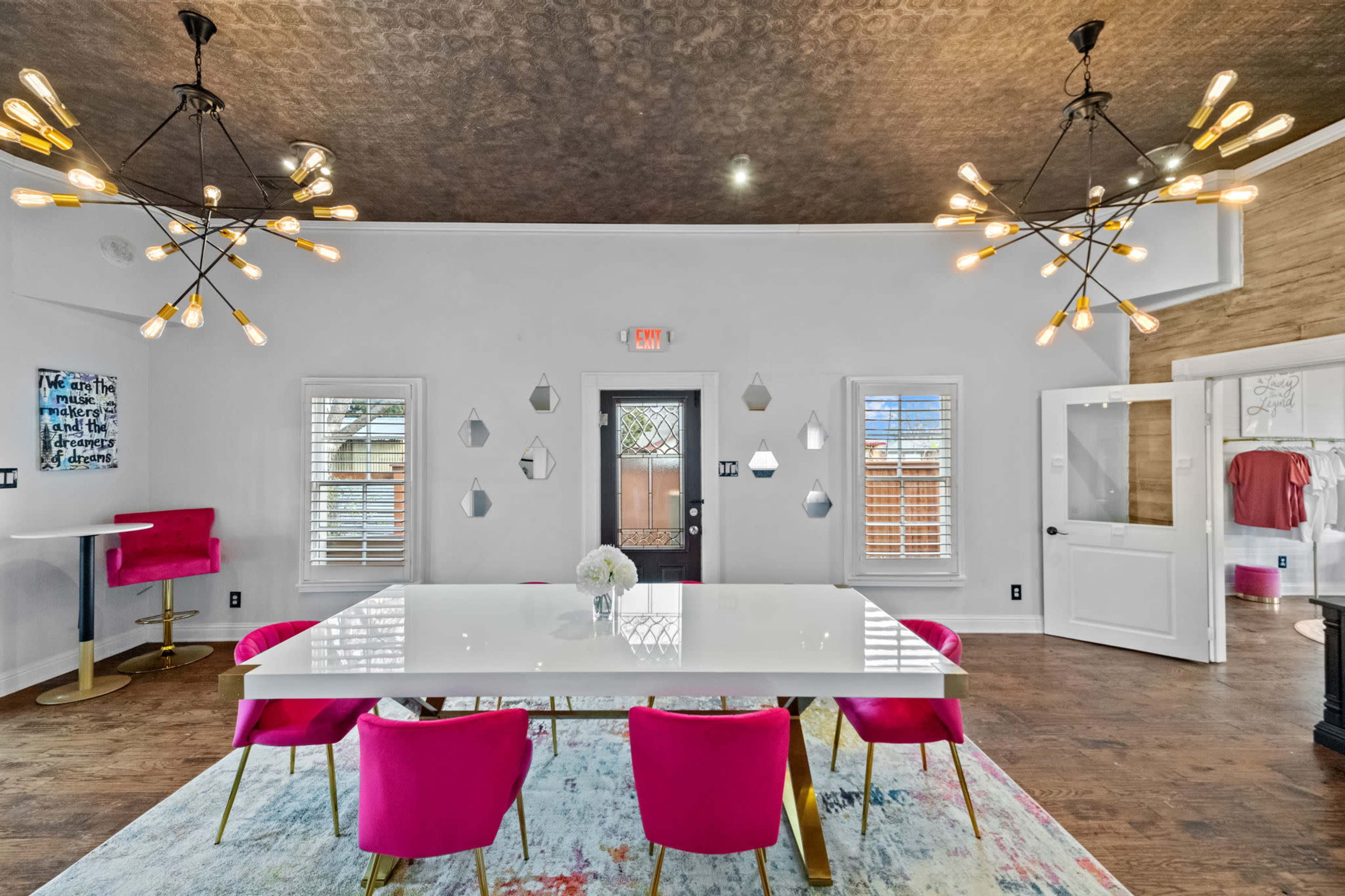 A modern dining area featuring a large white table surrounded by pink chairs, two unique light fixtures hanging from the ceiling, and decorative wall mirrors.