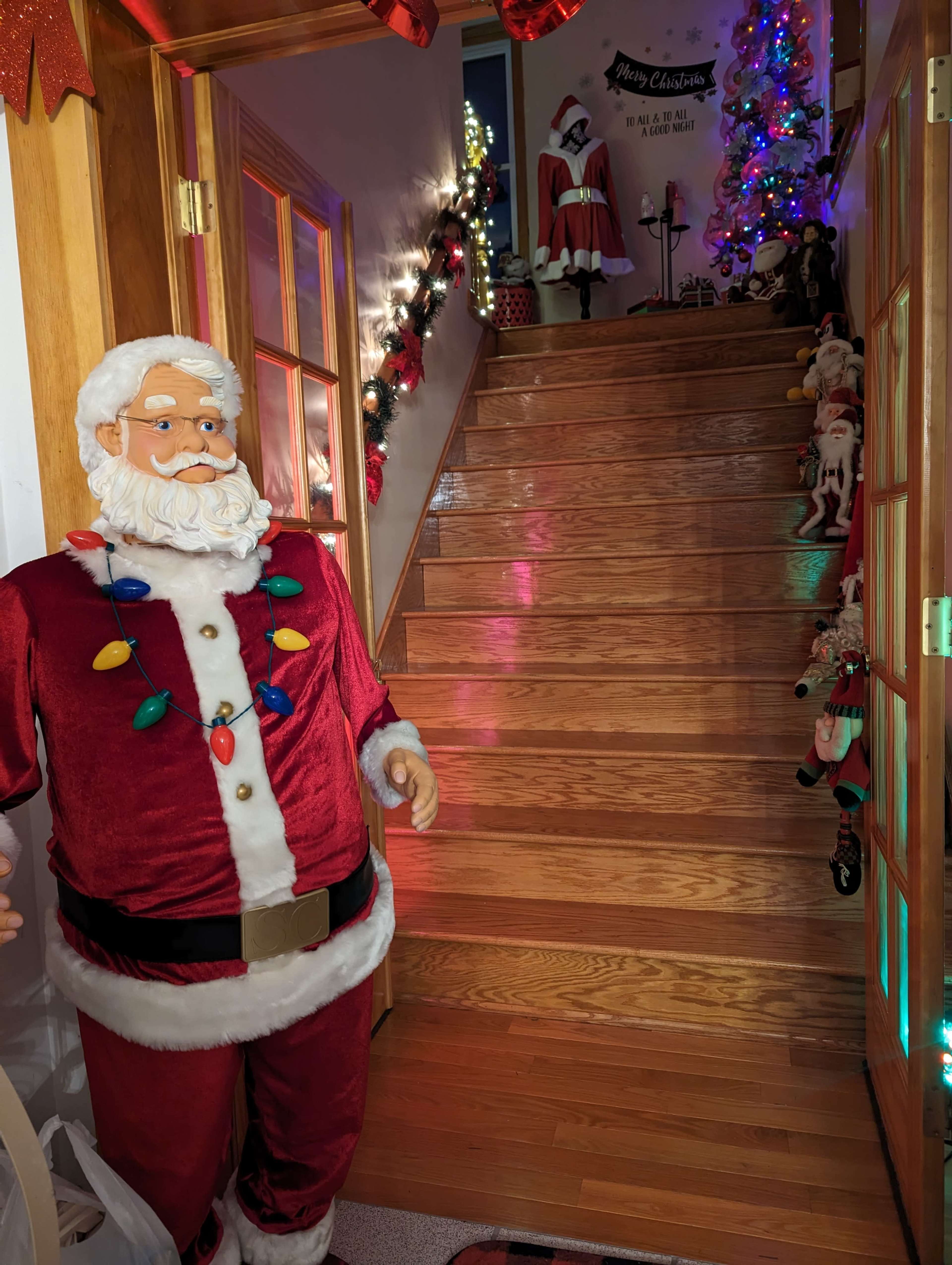 A life-sized Santa Claus figure adorned with festive lights stands next to a staircase decorated with Christmas ornaments and lights.