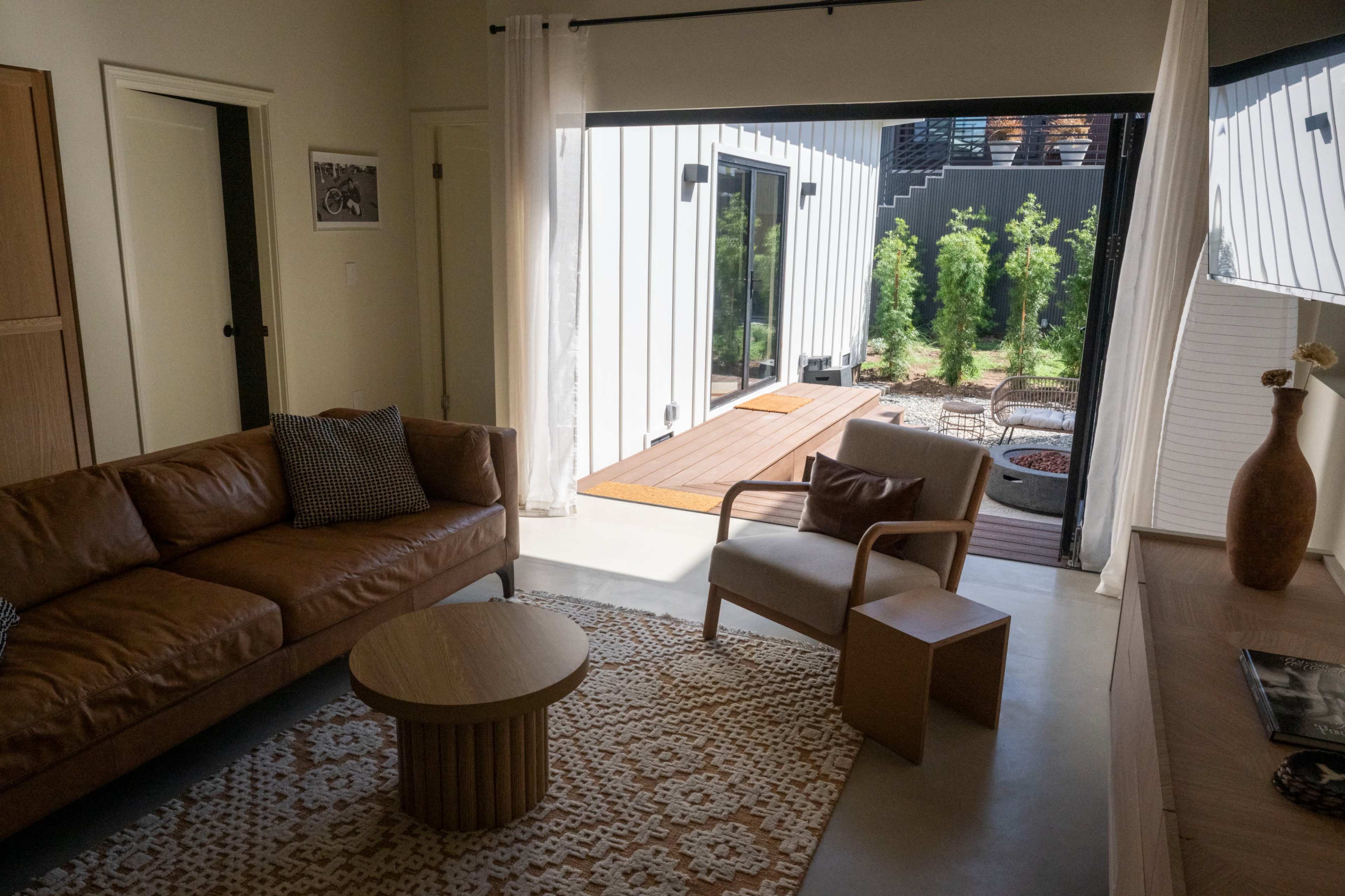 The image shows a modern living room with a brown leather sofa, a round wooden coffee table, and a gray armchair, leading to an outdoor space visible through a large sliding door.