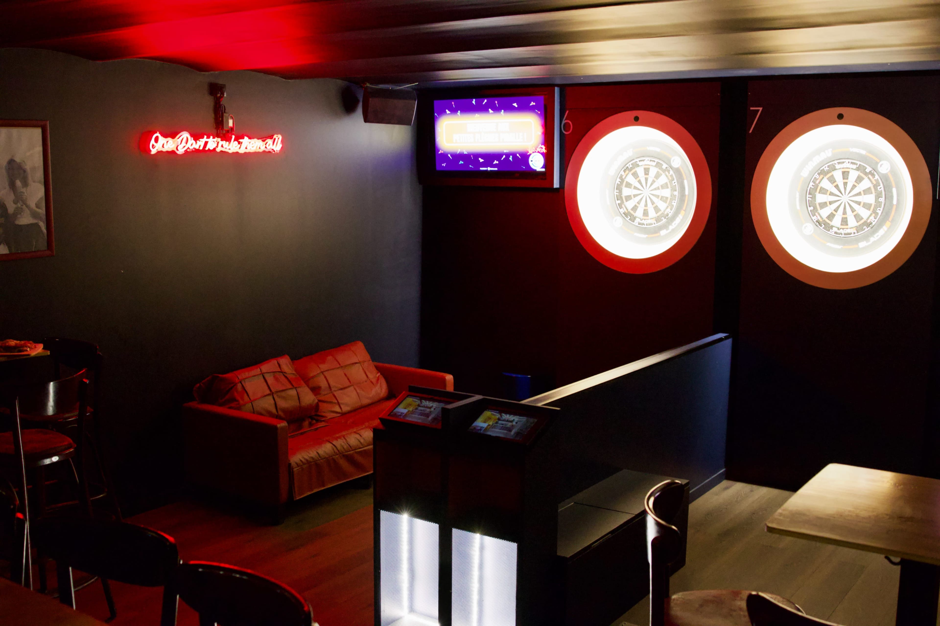 The image shows a dimly lit bar area featuring a red neon sign, a leather couch, and two dartboards illuminated on the wall.
