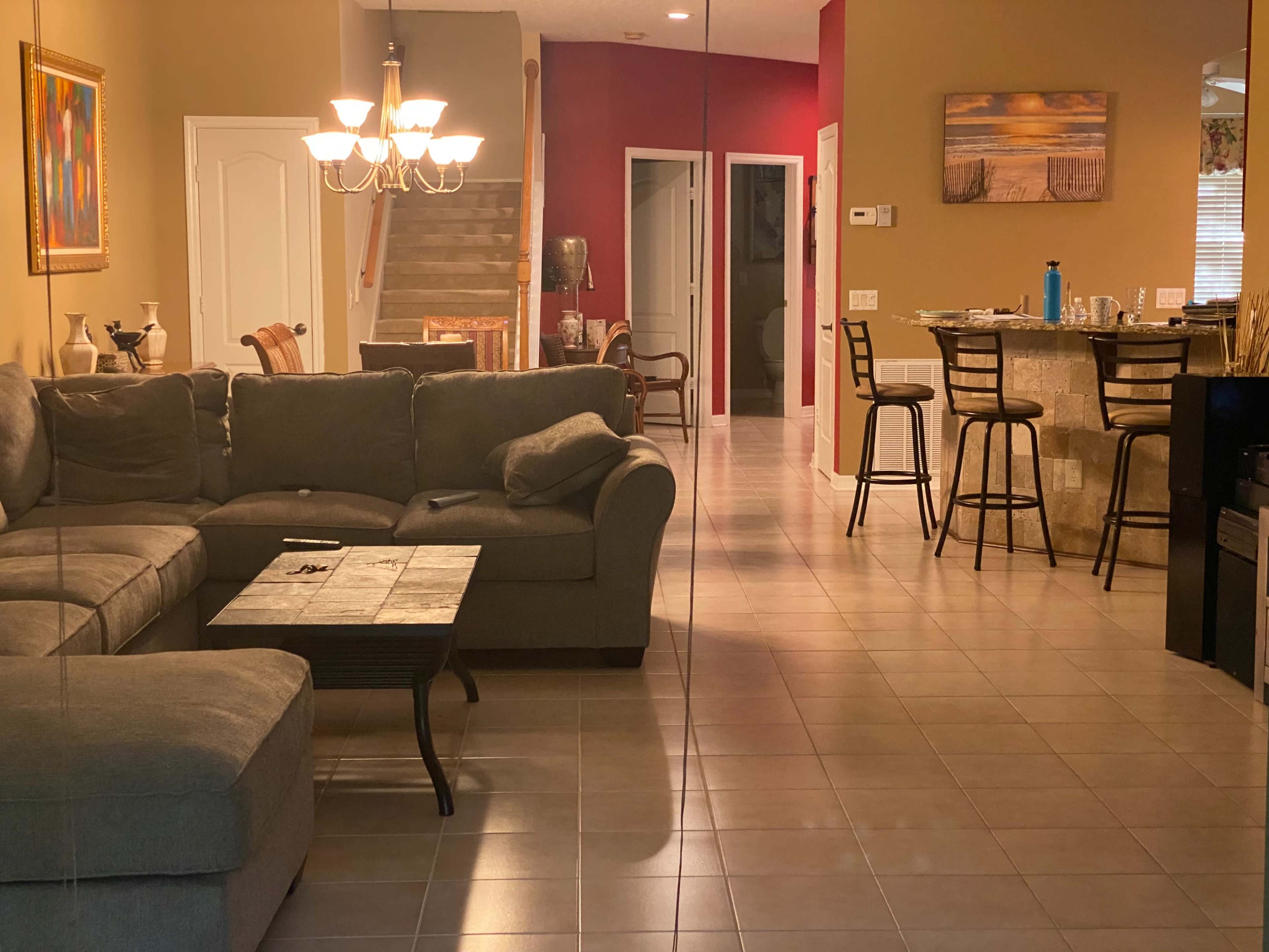A living room with a gray sectional sofa and a coffee table, leading to an open dining area and kitchen with bar stools.