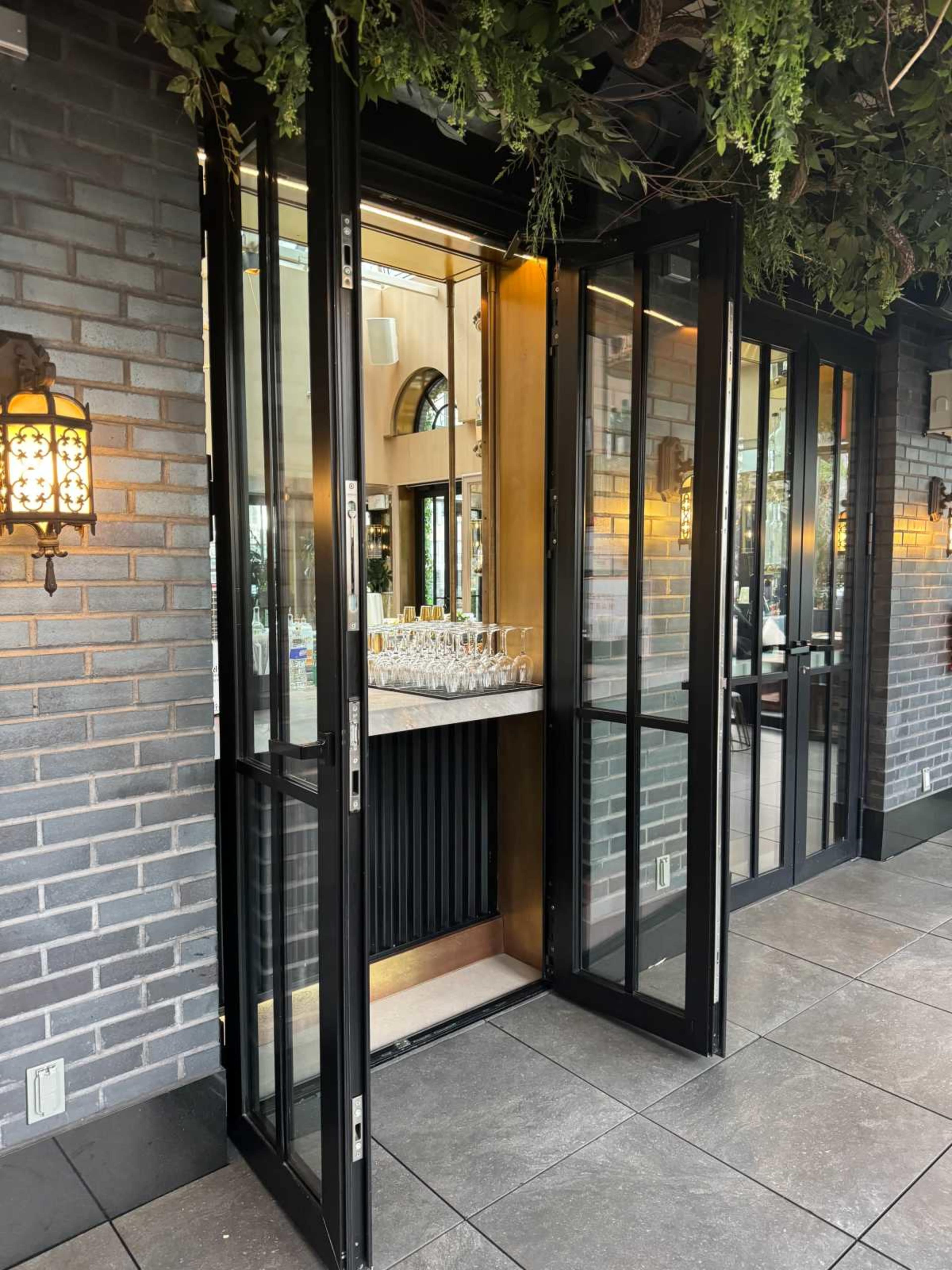 The image shows a set of glass doors that open into a restaurant, framed by a brick wall and illuminated by wall sconces.