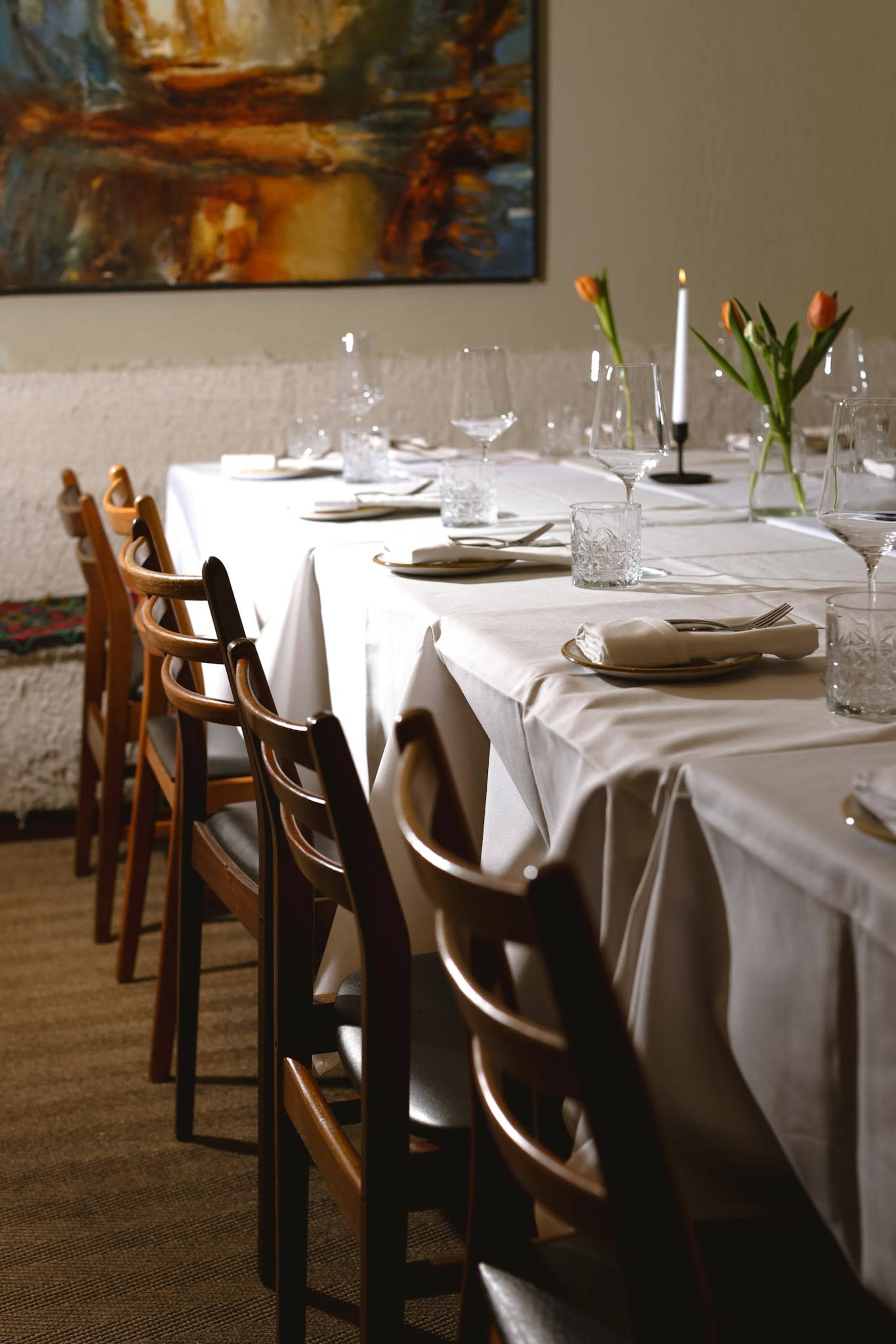 A neatly arranged dining table with white linens, glassware, and flowers, set in a warm-toned room with a colorful painting on the wall.