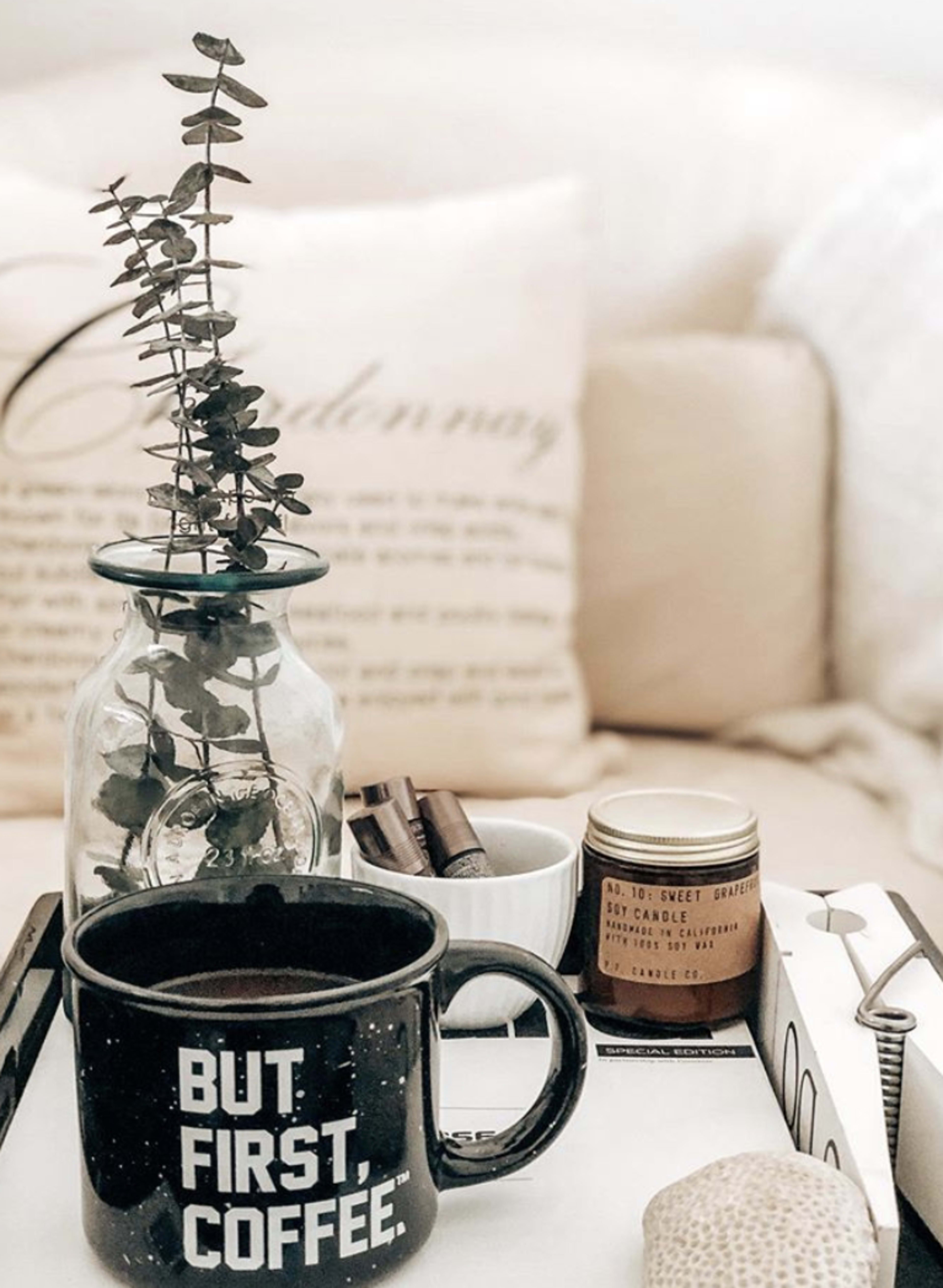 A black coffee mug with the phrase "BUT FIRST, COFFEE" sits on a tray alongside a small plant in a glass vase, a candle, and various writing utensils.