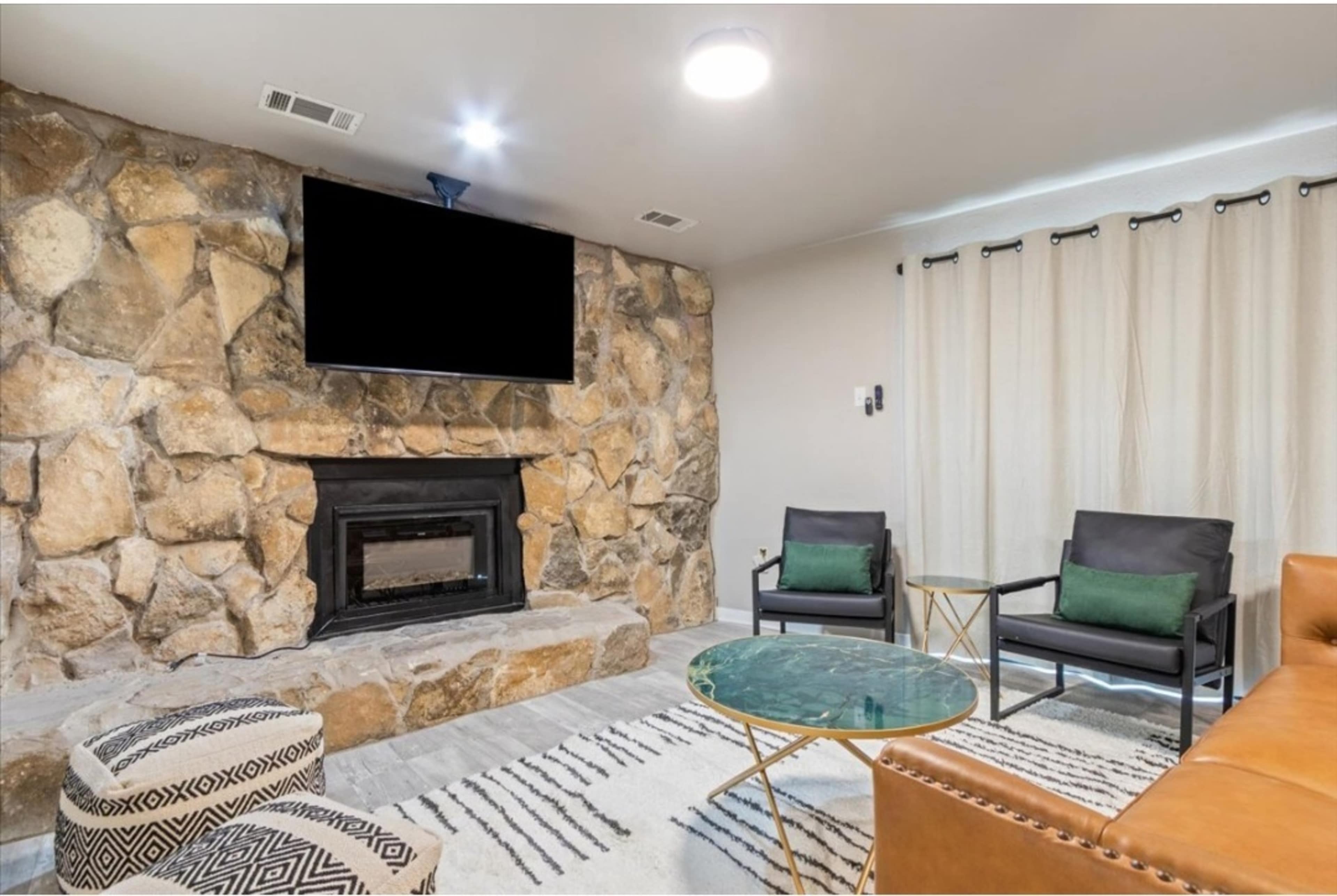 A living room with a stone fireplace, a large flat-screen TV mounted above, and modern furniture, including two black chairs and a brown leather sofa.
