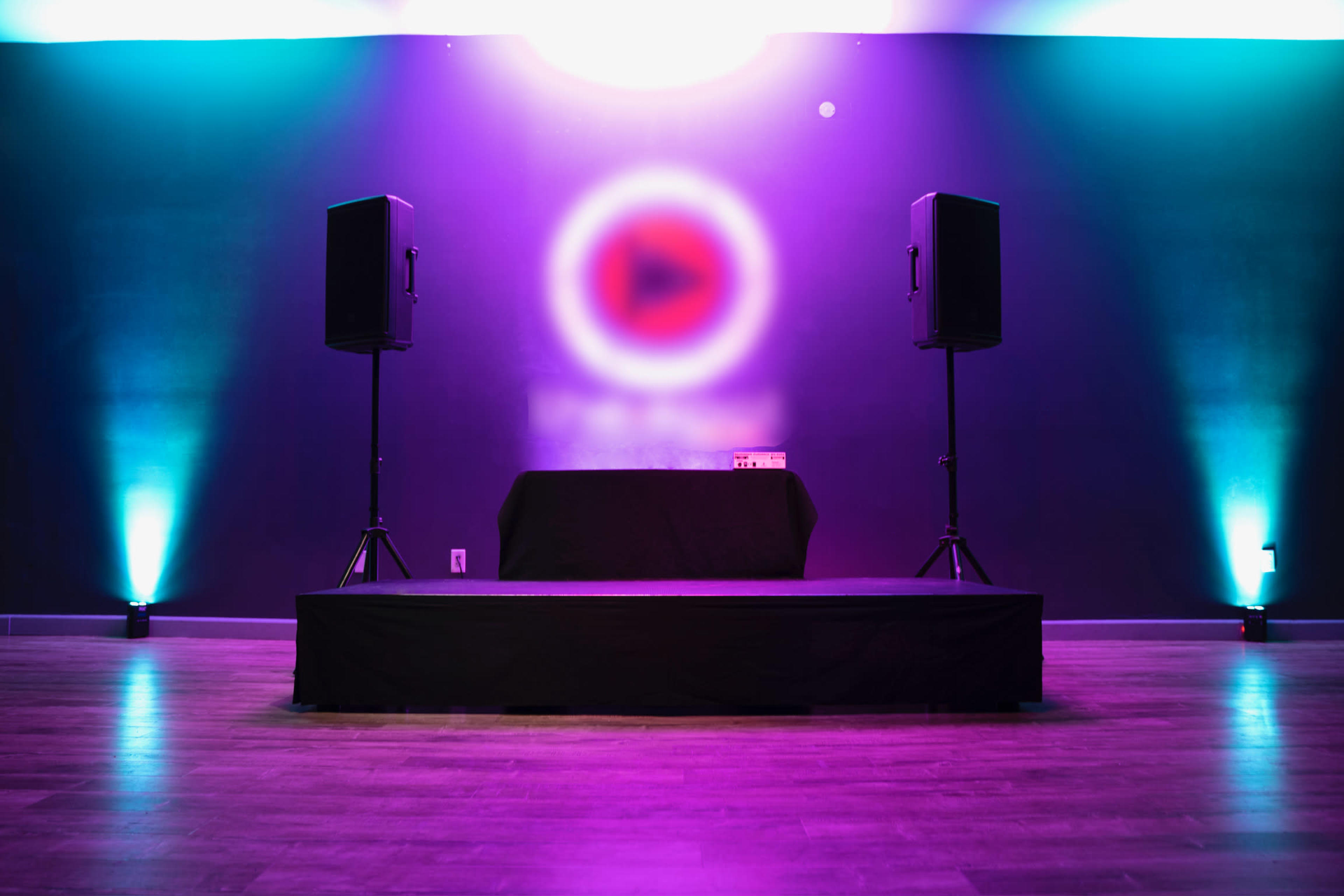A stage setup with two speakers flanking a central table, illuminated by purple and blue lighting against a dark wall.