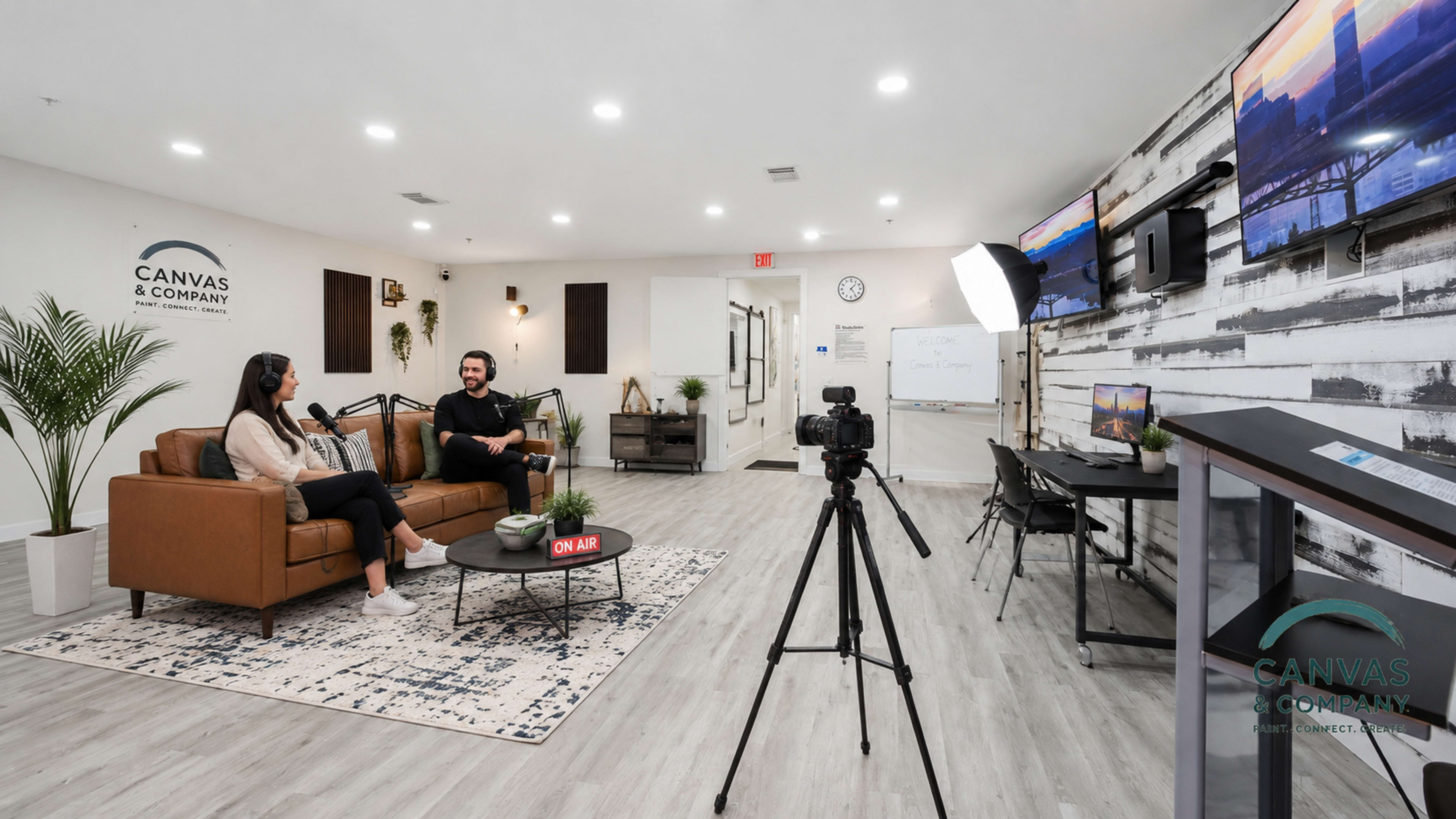 A modern studio setup features a couch with two people conversing, a camera on a tripod facing them, and televisions displaying content in a well-lit space.
