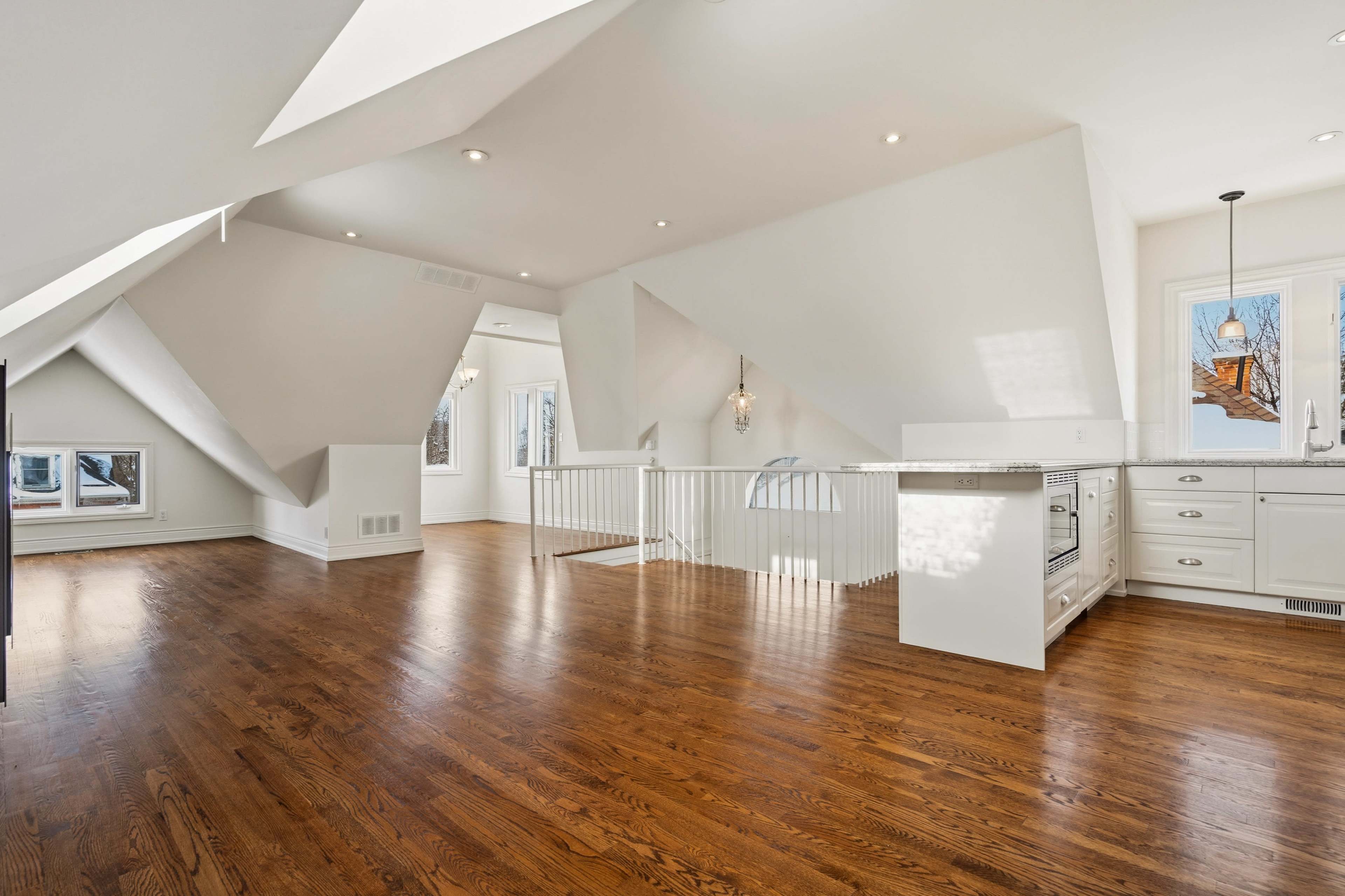 The image shows a spacious, minimalist interior of a room with sloped ceilings, wooden flooring, and large windows allowing natural light to fill the space.