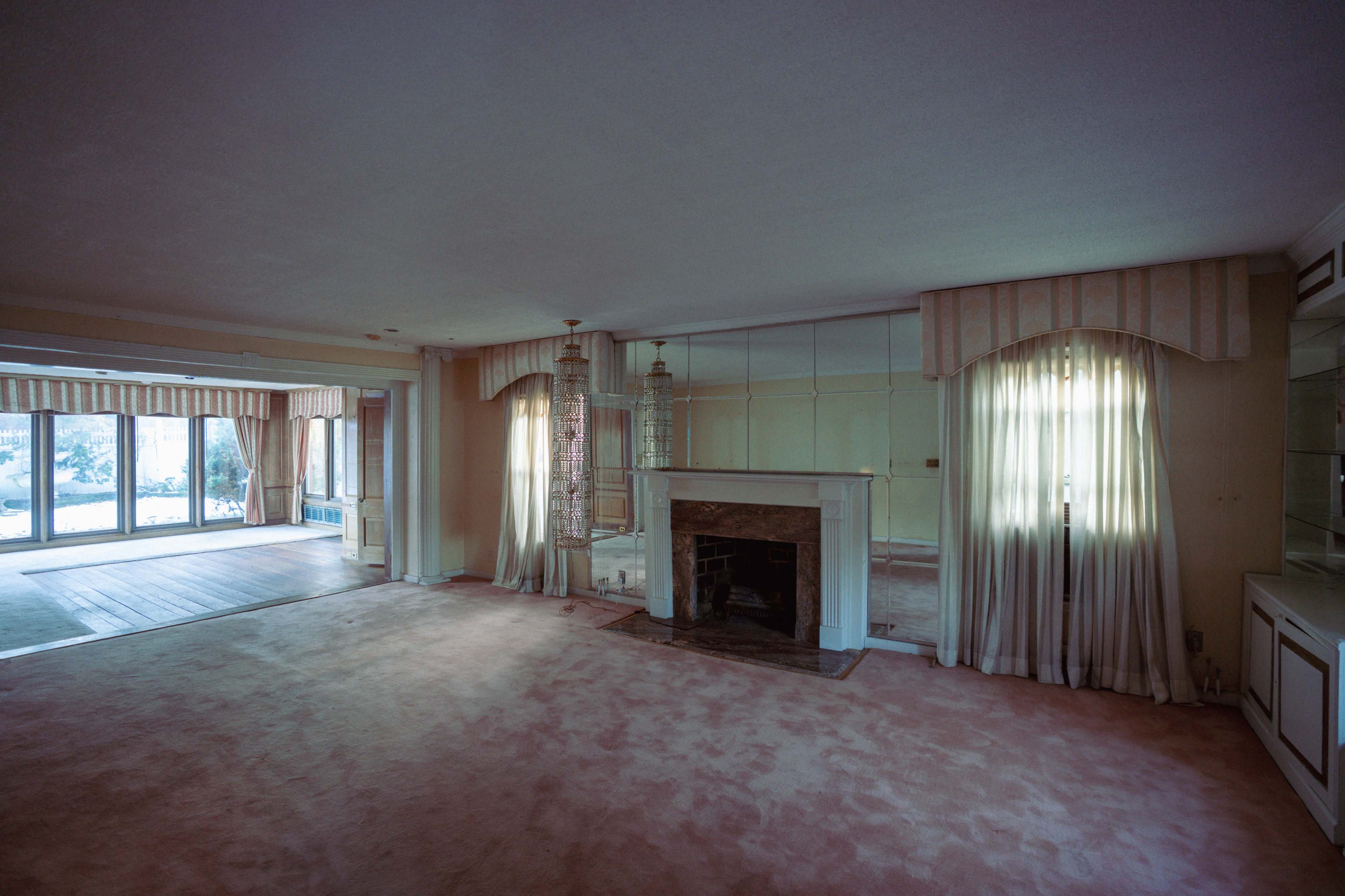 The image shows a spacious, empty living room with a fireplace, large windows, and mirrored walls.