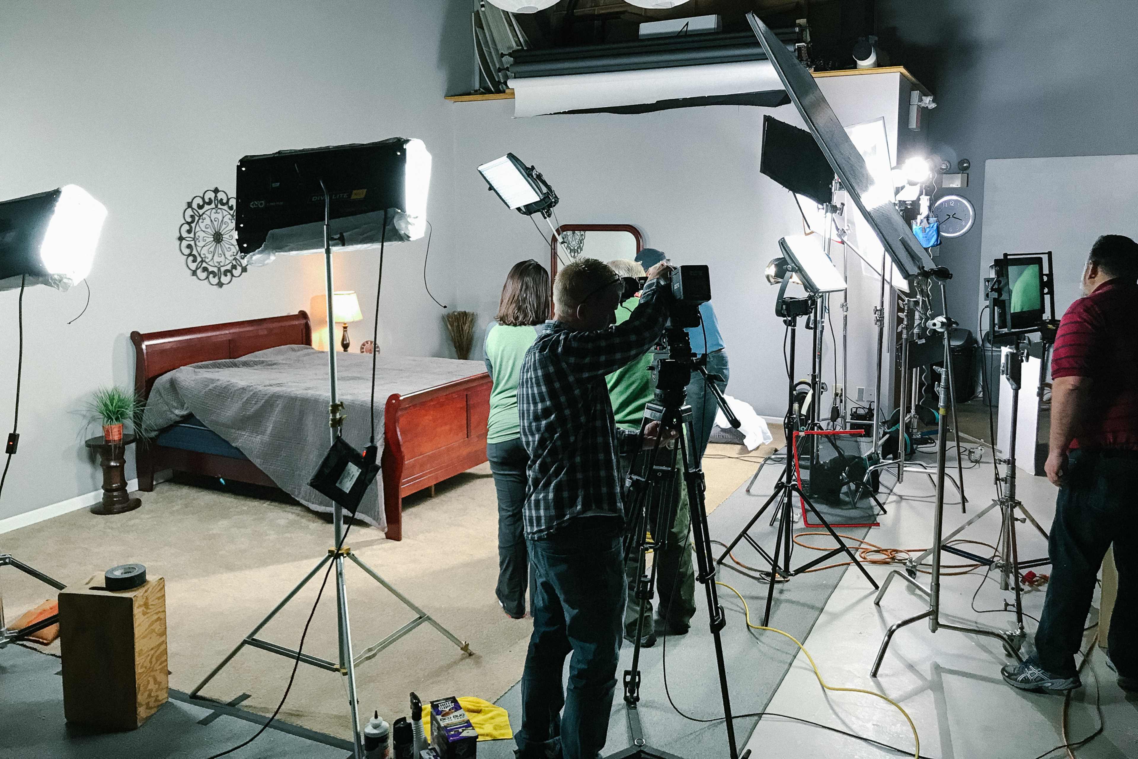 A film crew sets up equipment around a bedroom scene with a wooden bed, lighting rigs, and cameras.