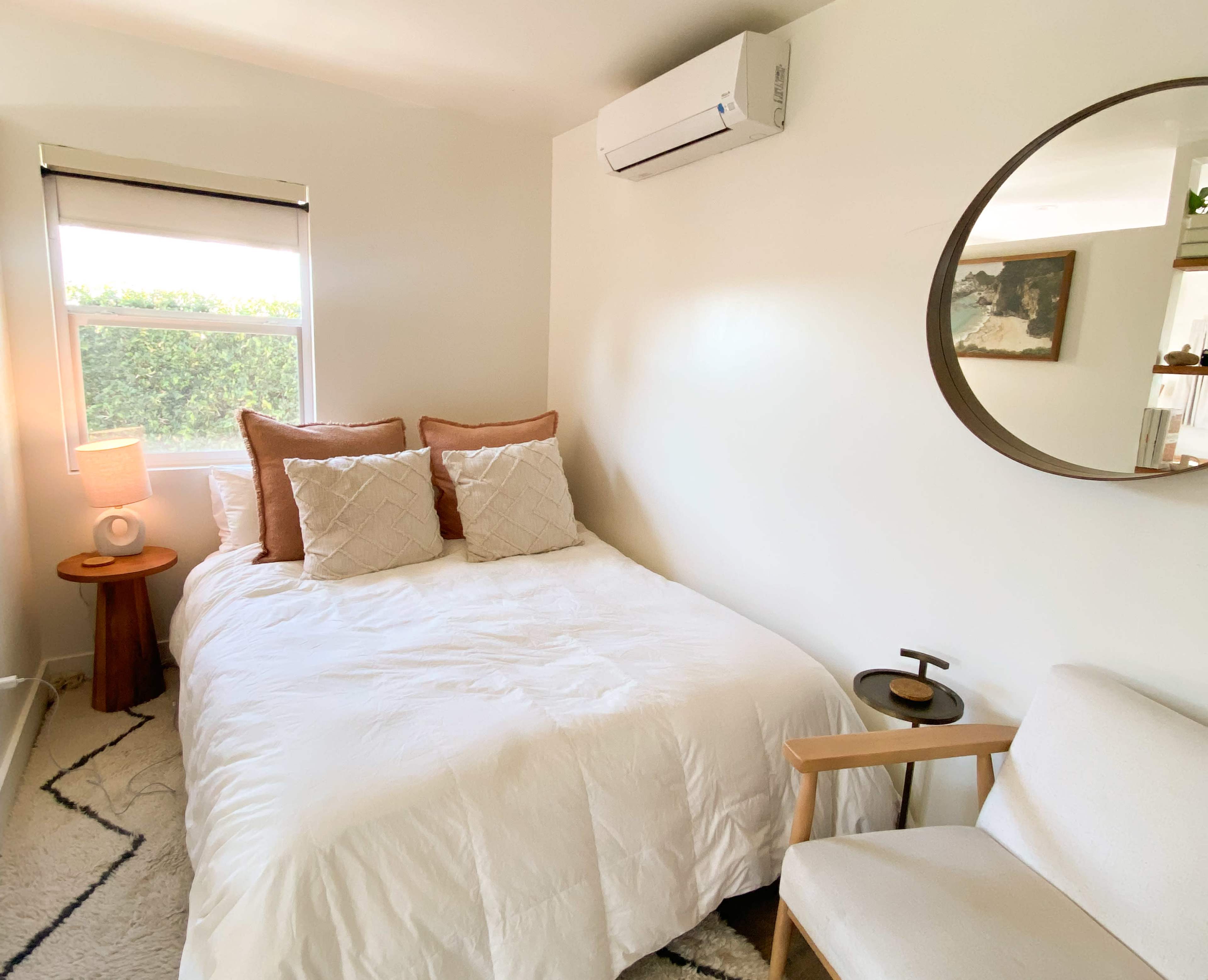 The image shows a well-lit bedroom featuring a white bed with decorative pillows, a small side table with a lamp, and a cozy chair next to a round mirror on a neutral-colored wall.