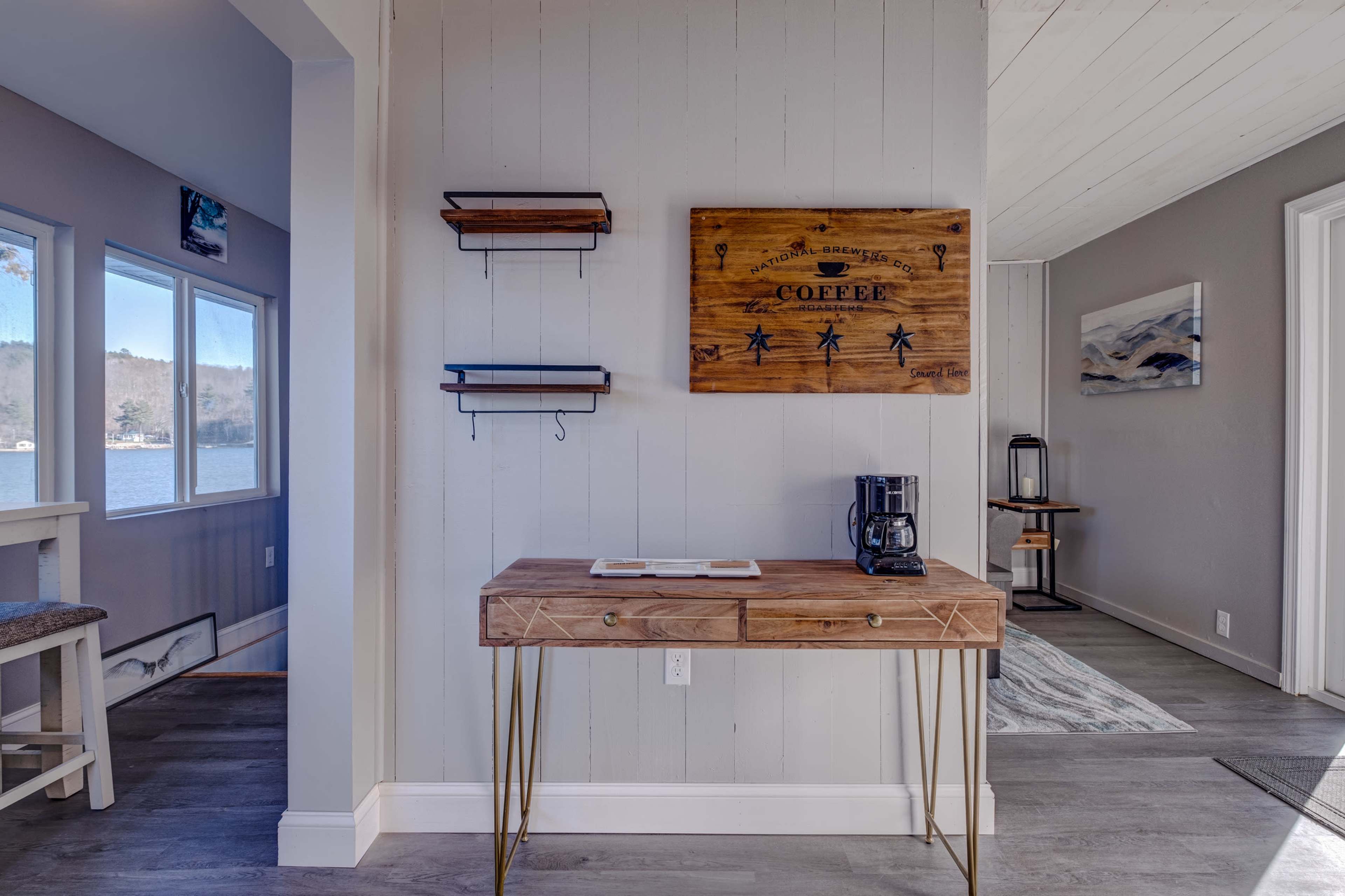 A wooden table with a coffee maker and a decorative sign on the wall, set in a room with large windows overlooking a lake.