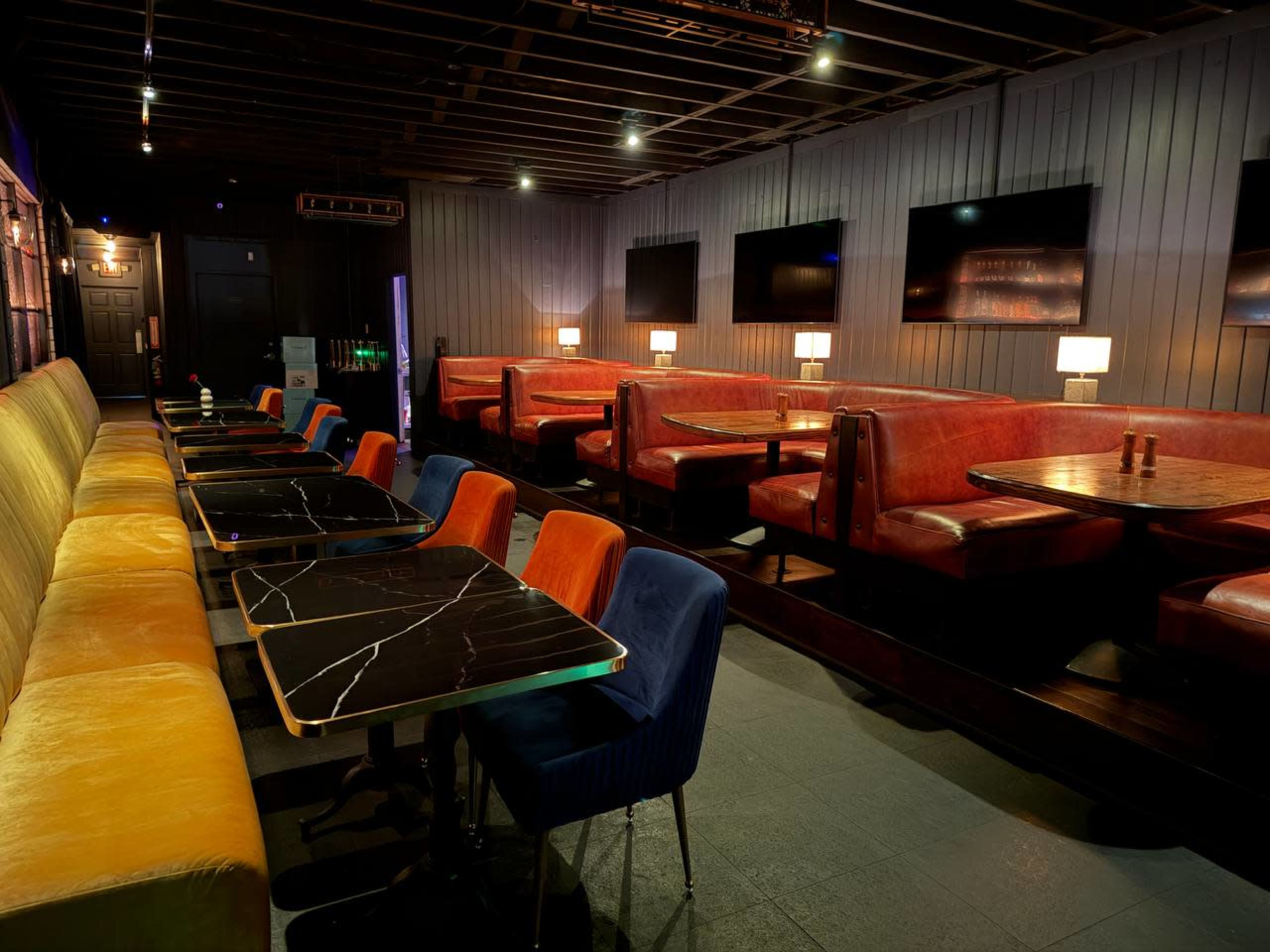 The image shows a modern lounge or bar with colorful seating arrangements, dark tables, and several flat-screen TVs mounted on the walls.