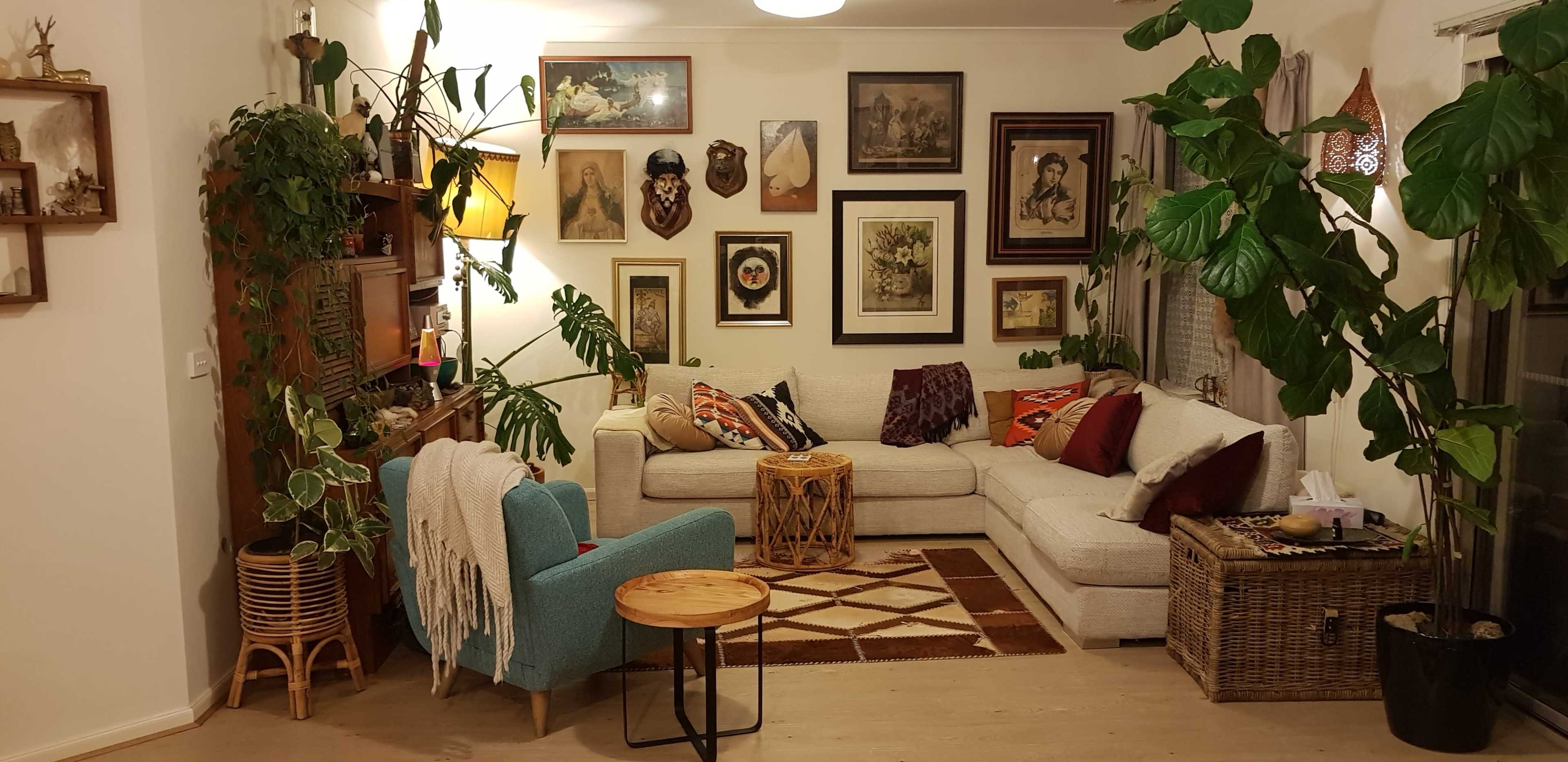 A cozy living room features a large sectional sofa, various framed artworks on the walls, an array of indoor plants, and a mix of decorative furniture.
