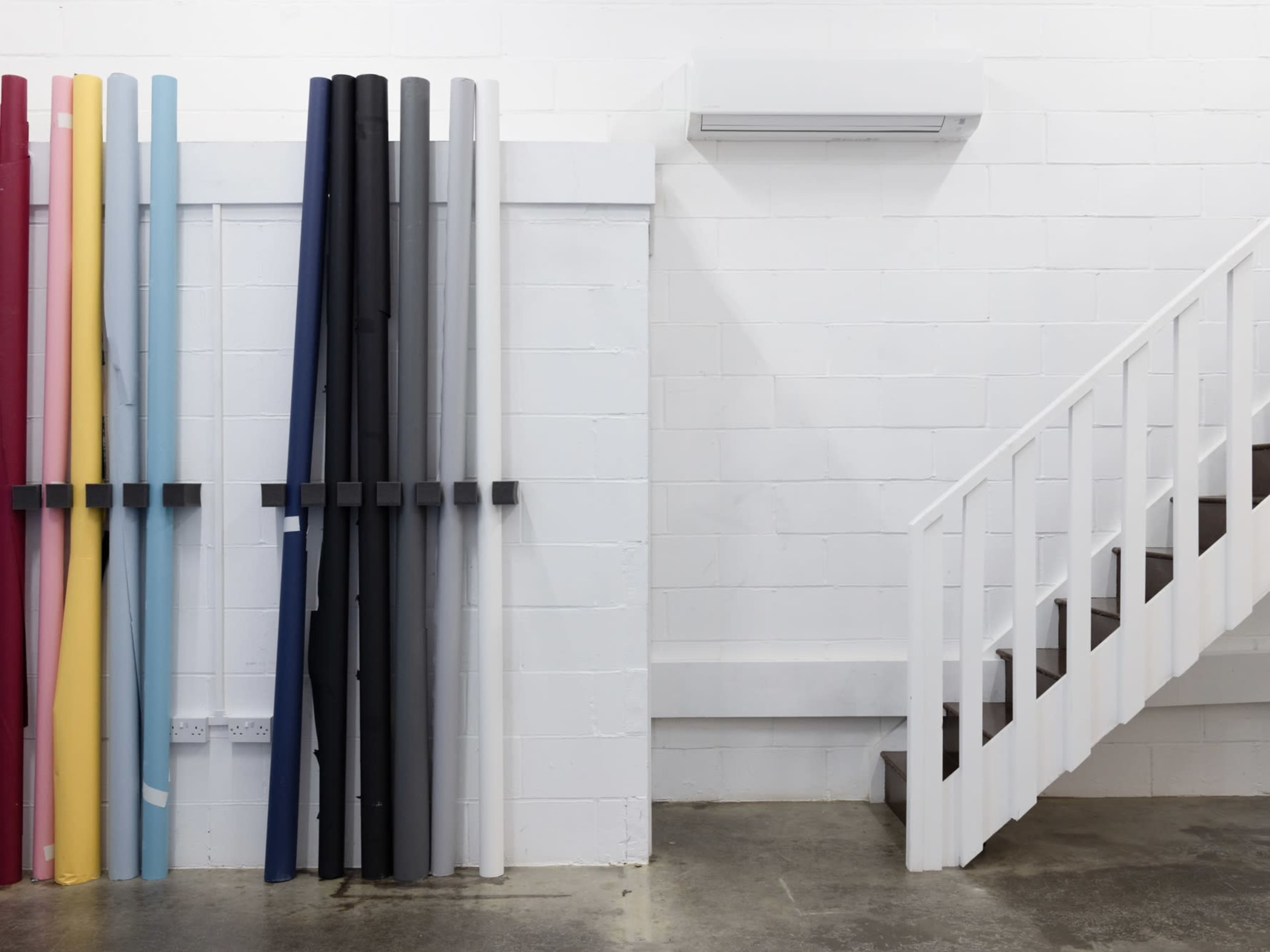 A wall with several rolls of colored material neatly arranged beside a white staircase in a minimalist interior space.