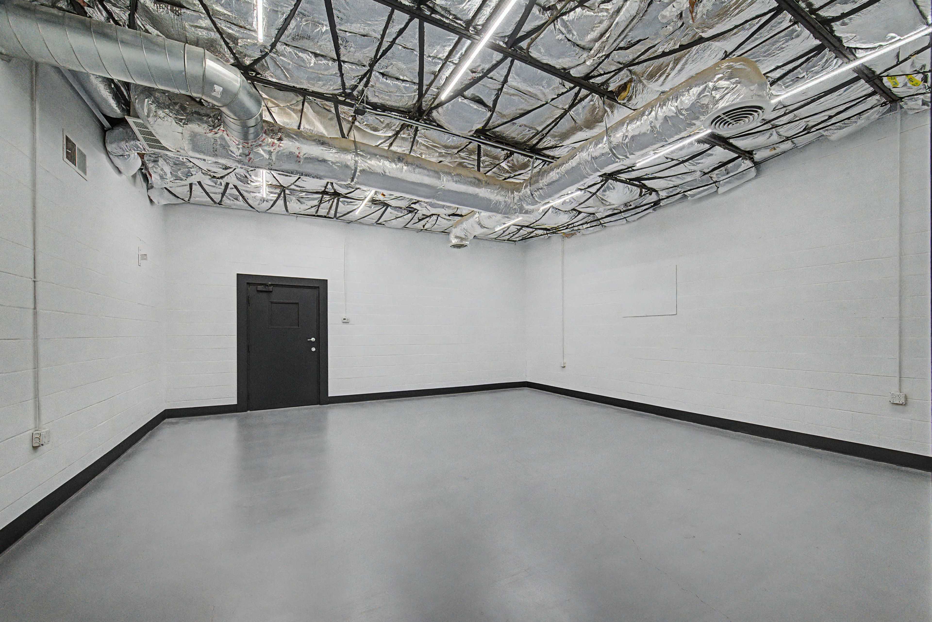 The image shows an empty room with a gray floor, white cinder block walls, and a metal ducted ceiling.
