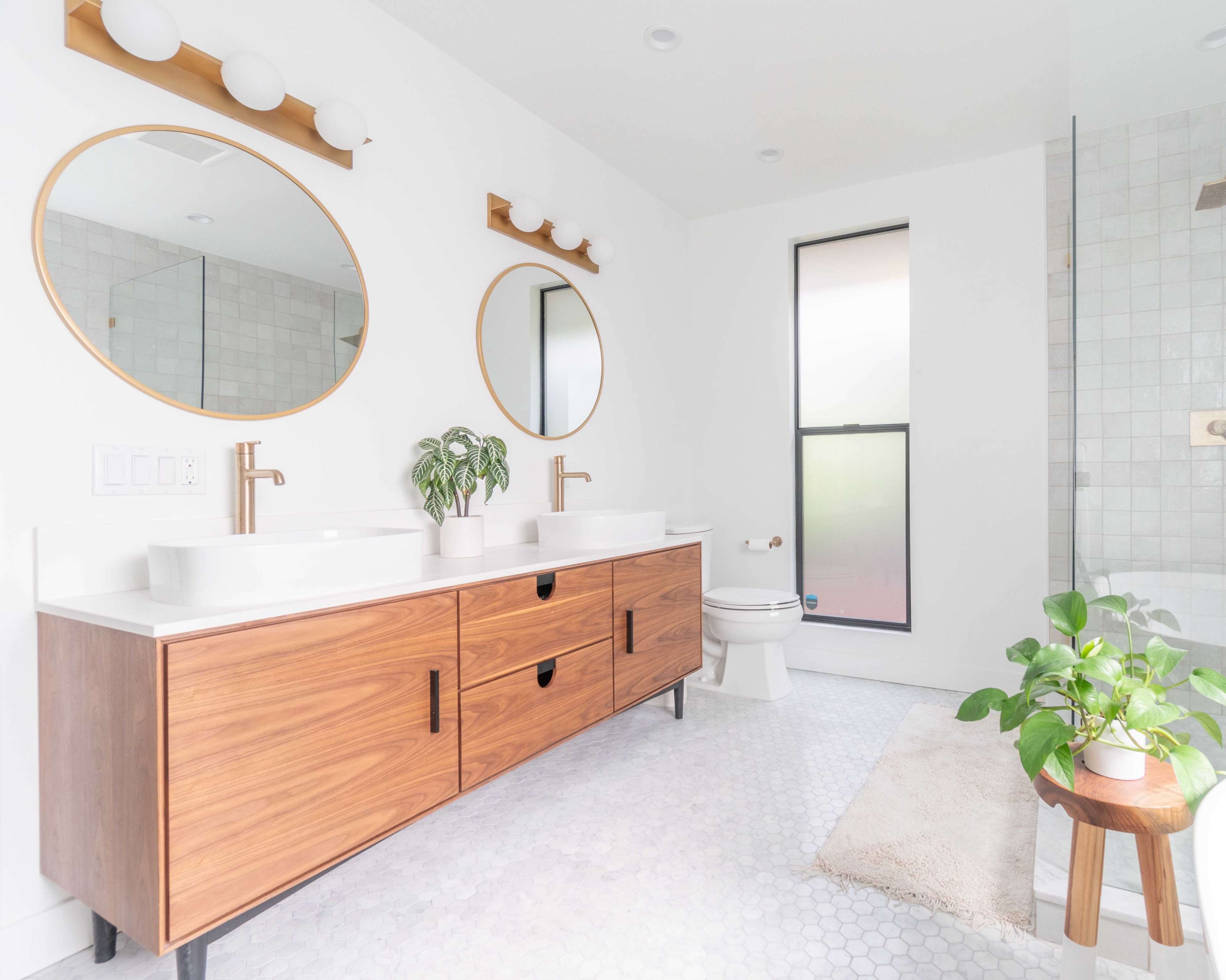 The image shows a modern bathroom featuring a double vanity with a wooden cabinet, two round mirrors, a glass shower, and a potted plant on a stool.