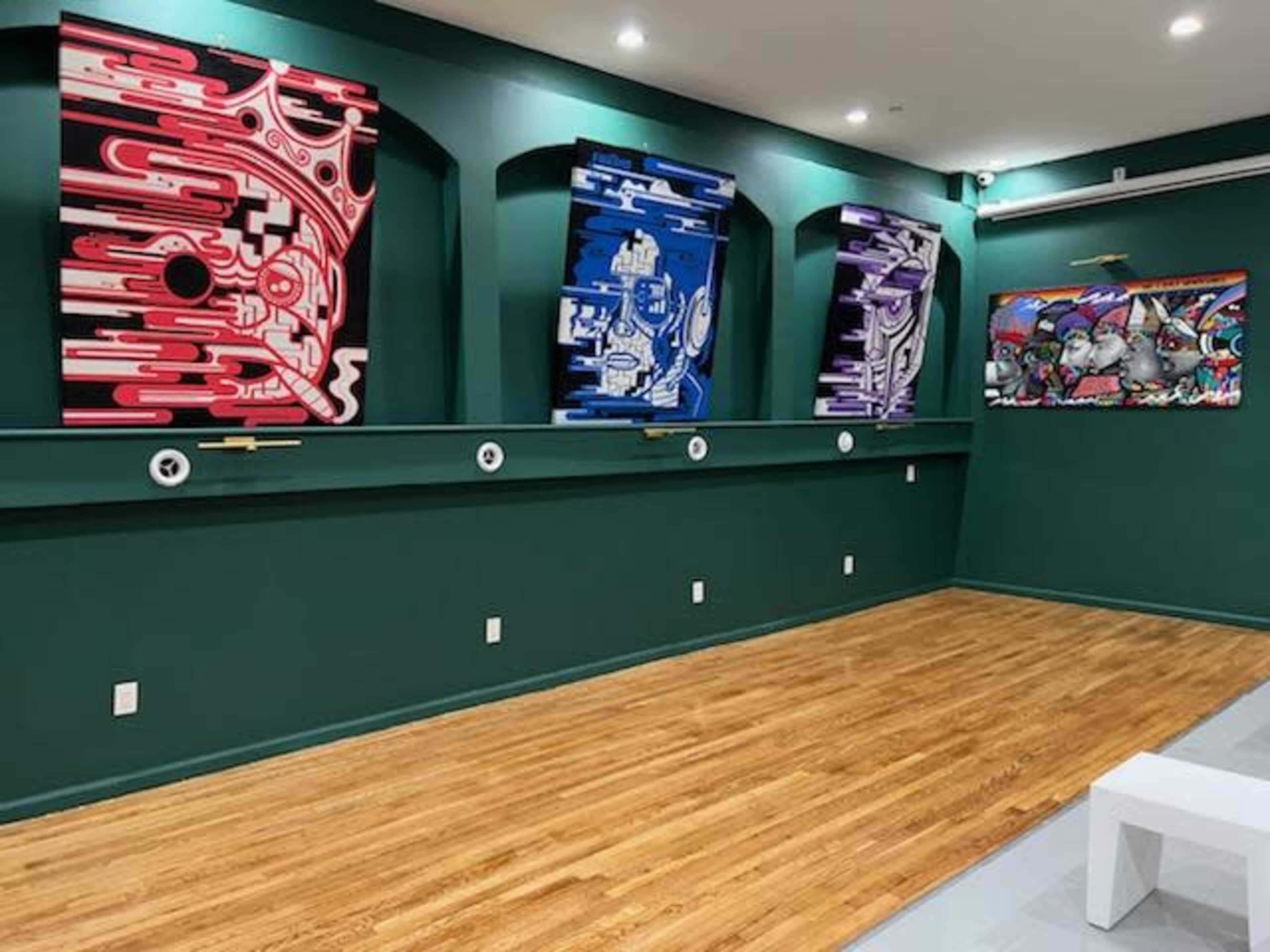 An art gallery wall featuring four colorful abstract artworks on a green backdrop, with wooden flooring and a minimalist display setup.