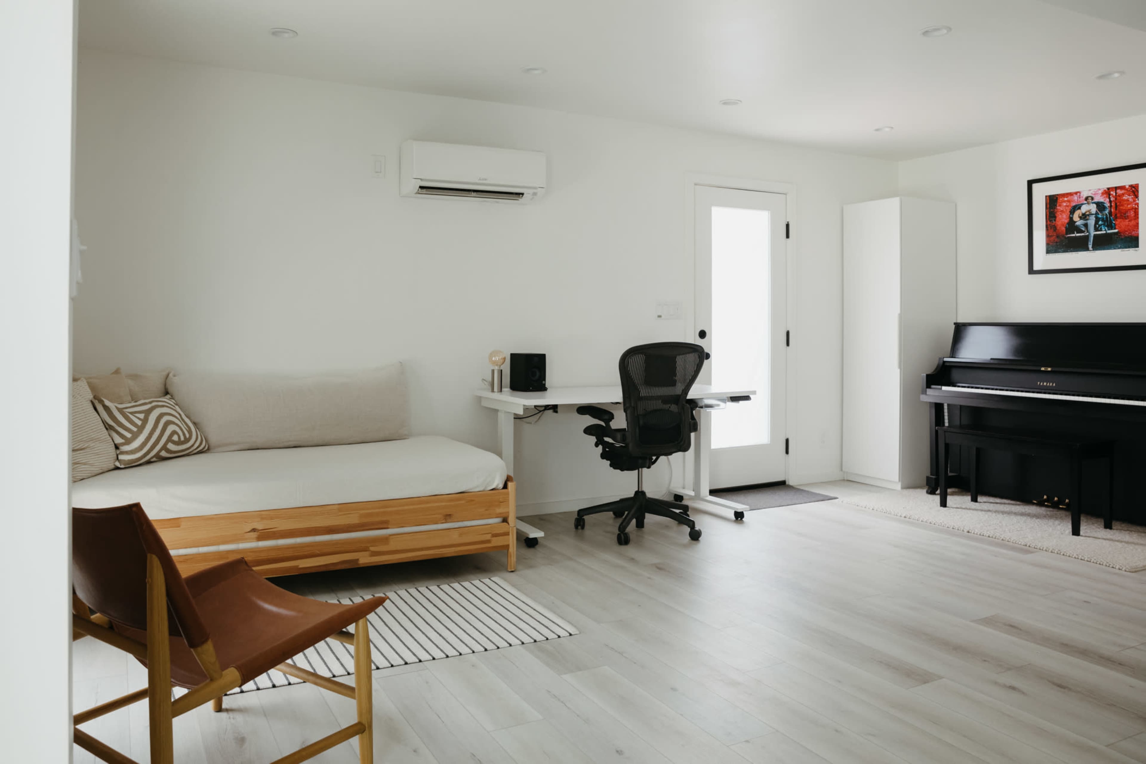 A minimalist room featuring a daybed, a desk with an office chair, and a piano, along with light-colored walls and wooden flooring.