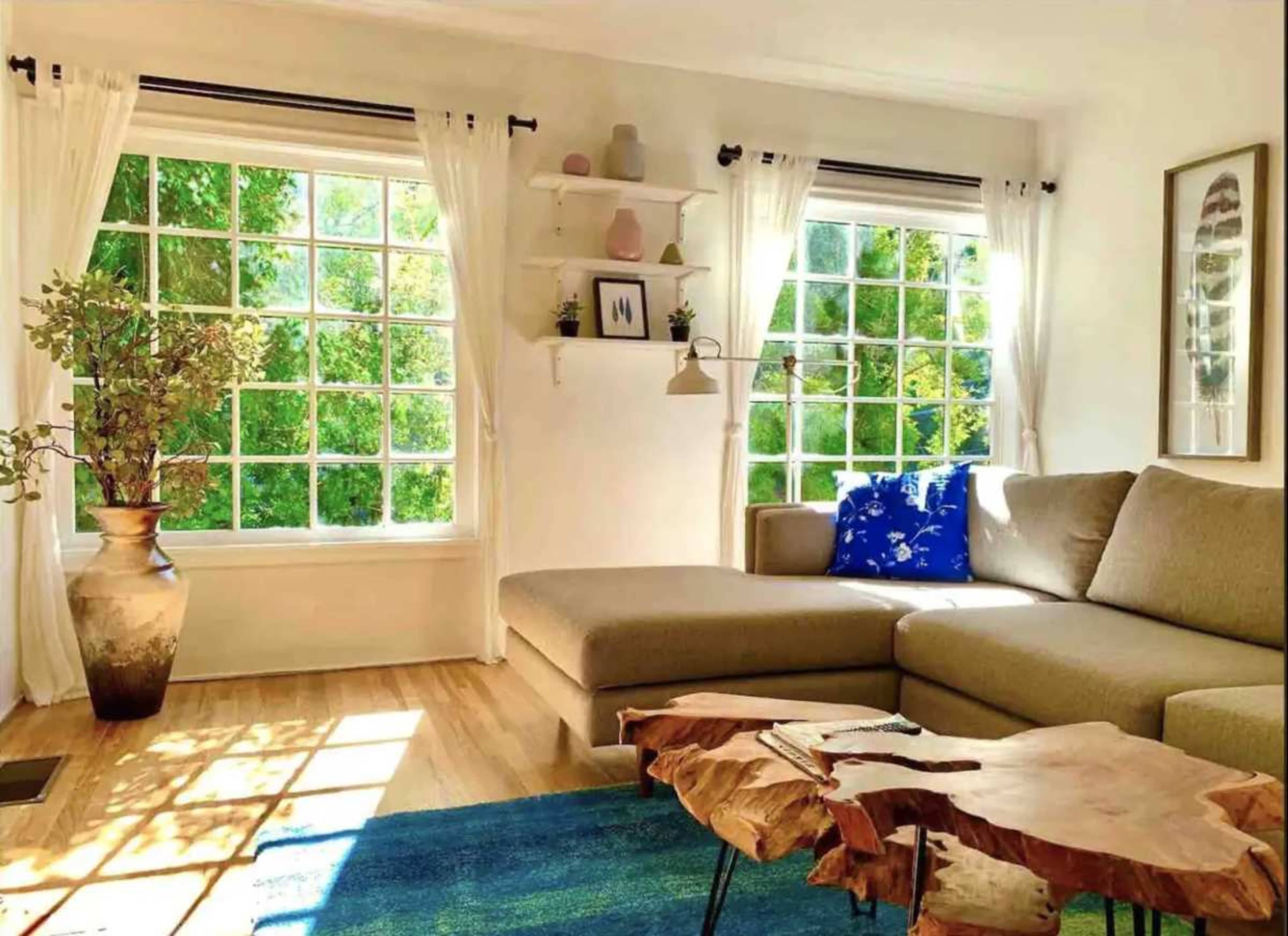 A bright living room features a sectional sofa, a wooden coffee table, and large windows with white curtains allowing natural light to fill the space.